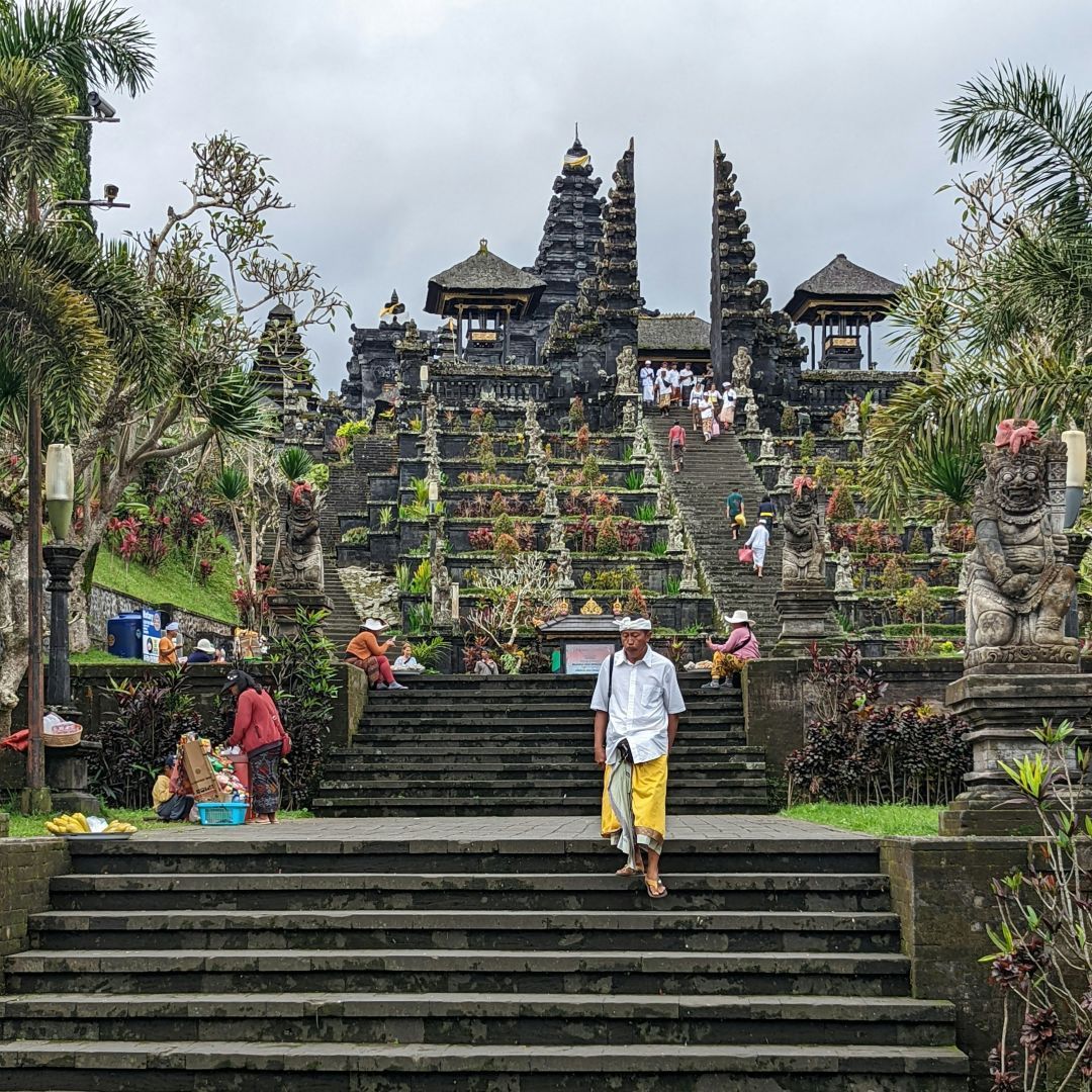 Ein Mann in weißem Hemd und gelbem Sarong schreitet die Steinstufen eines reich verzierten, mehrstufigen Tempels hinab, der von Gärten umgeben ist.