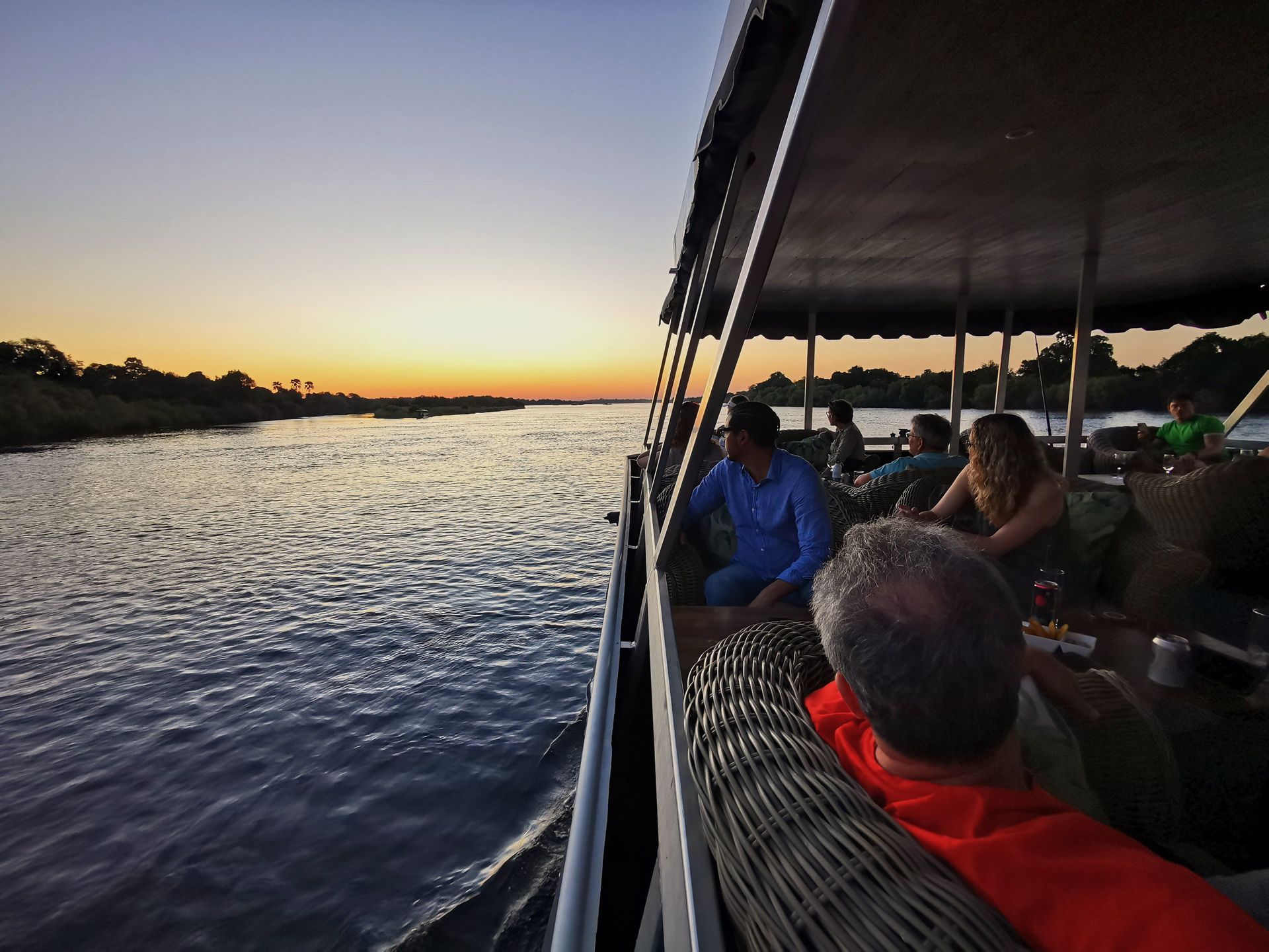 Un gruppo WeRoad in viaggio si rilassa sul ponte coperto di una barca ammirando il tramonto su un ampio fiume.