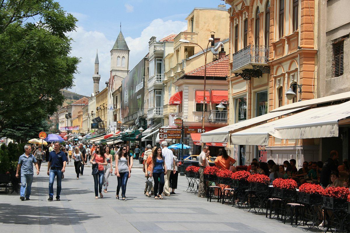 An einem sonnigen Tag schlendern Menschen durch eine belebte Fußgängerzone, gesäumt von Geschäften und Cafés mit Außenterrassen.