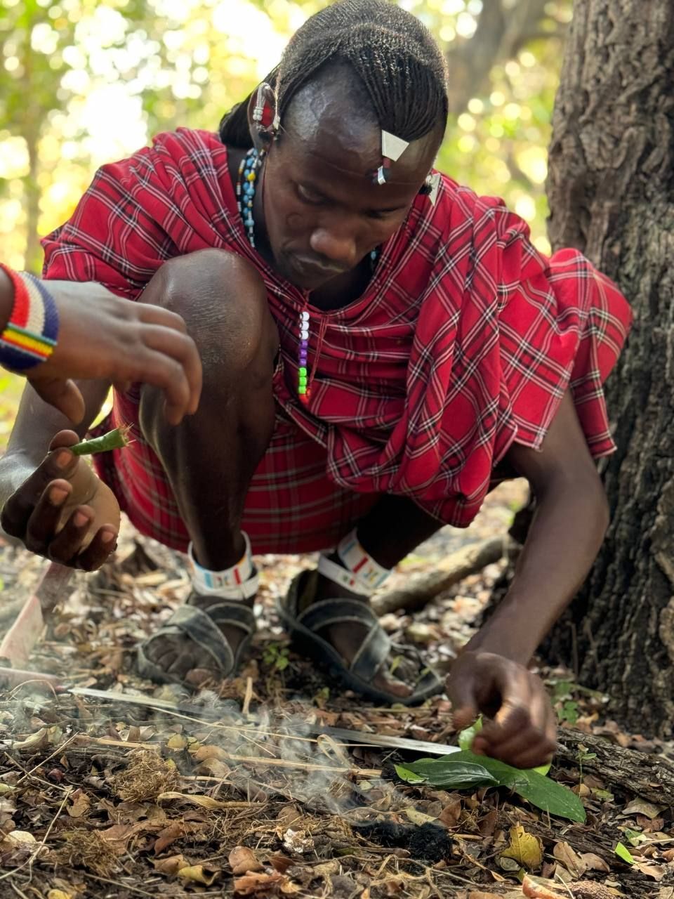 Un uomo con abiti tradizionali Maasai rossi è accovacciato sul suolo della foresta, intento a curare un piccolo fuoco fumante con foglie.