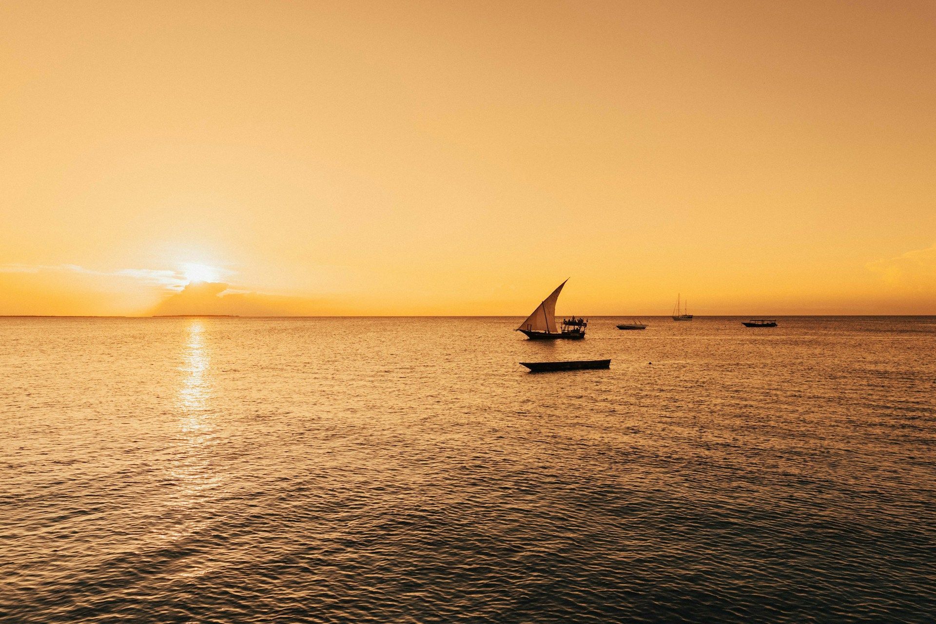 Un tradizionale veliero dhow e altre barche sull'oceano durante un tramonto dorato, con il sole che si riflette sull'acqua.