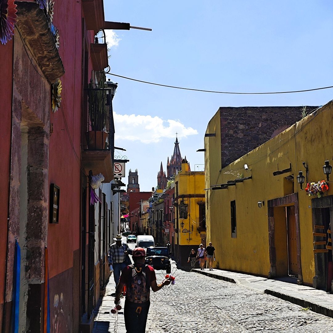 Ein Straßenhändler läuft durch eine enge, von farbenfrohen Gebäuden gesäumte Kopfsteinpflasterstraße, im Hintergrund ragen Kirchtürme auf.