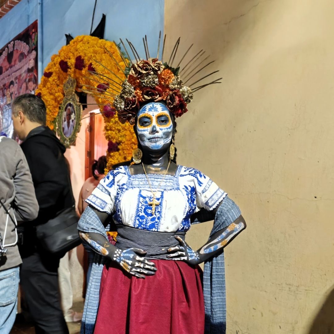 Eine Person in einem Catrina-Kostüm mit Totenkopf-Schminke und Blumenkopfschmuck posiert mit den Händen in den Hüften.