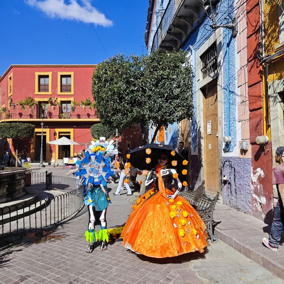 Zwei Personen in aufwendigen Tag der Toten Skelett-Kostümen stehen auf einer Kopfsteinpflasterstraße vor farbenfrohen Gebäuden.