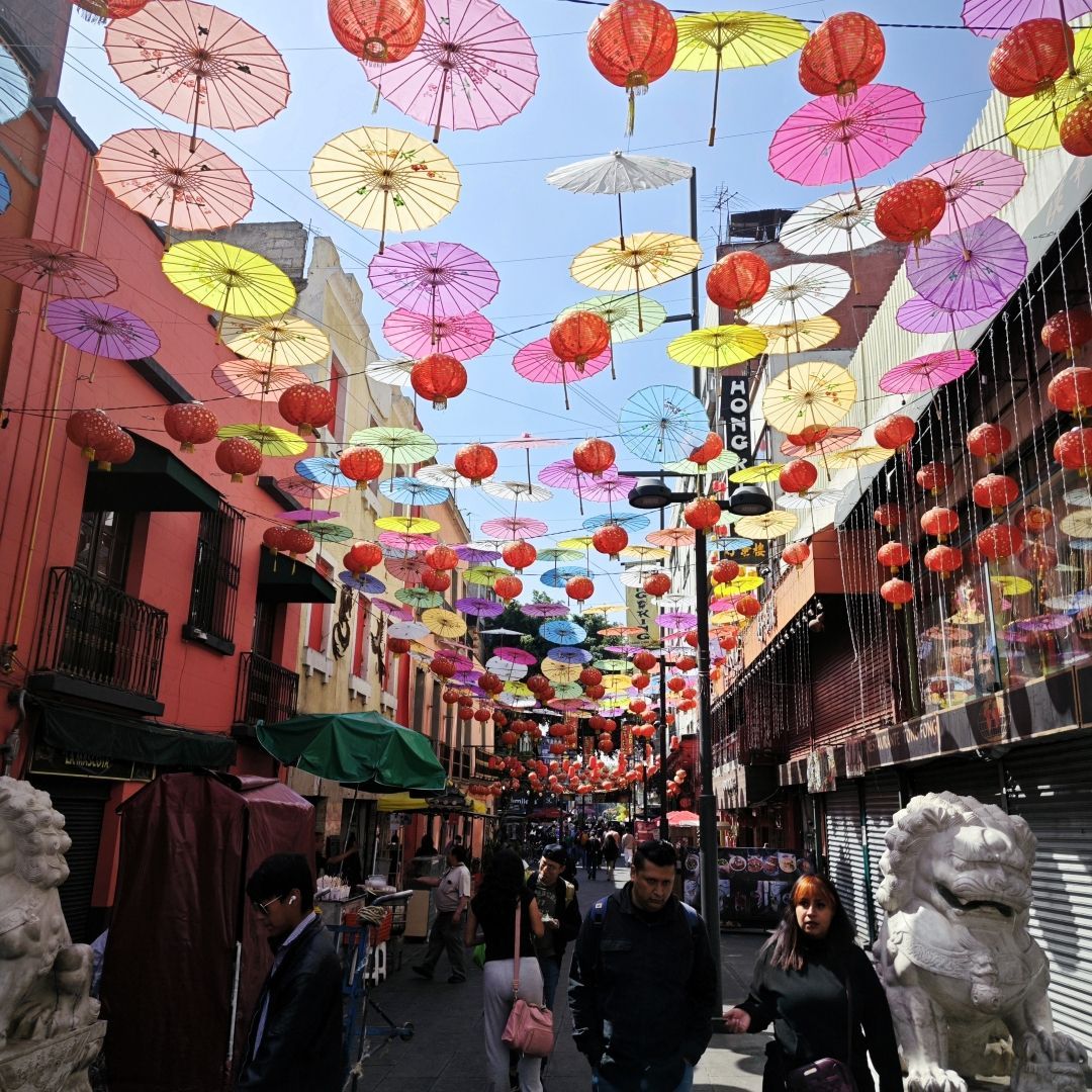 Bunte Papierschirme und rote Laternen hängen über einer belebten Stadtstraße, wo Menschen zwischen Gebäuden spazieren.