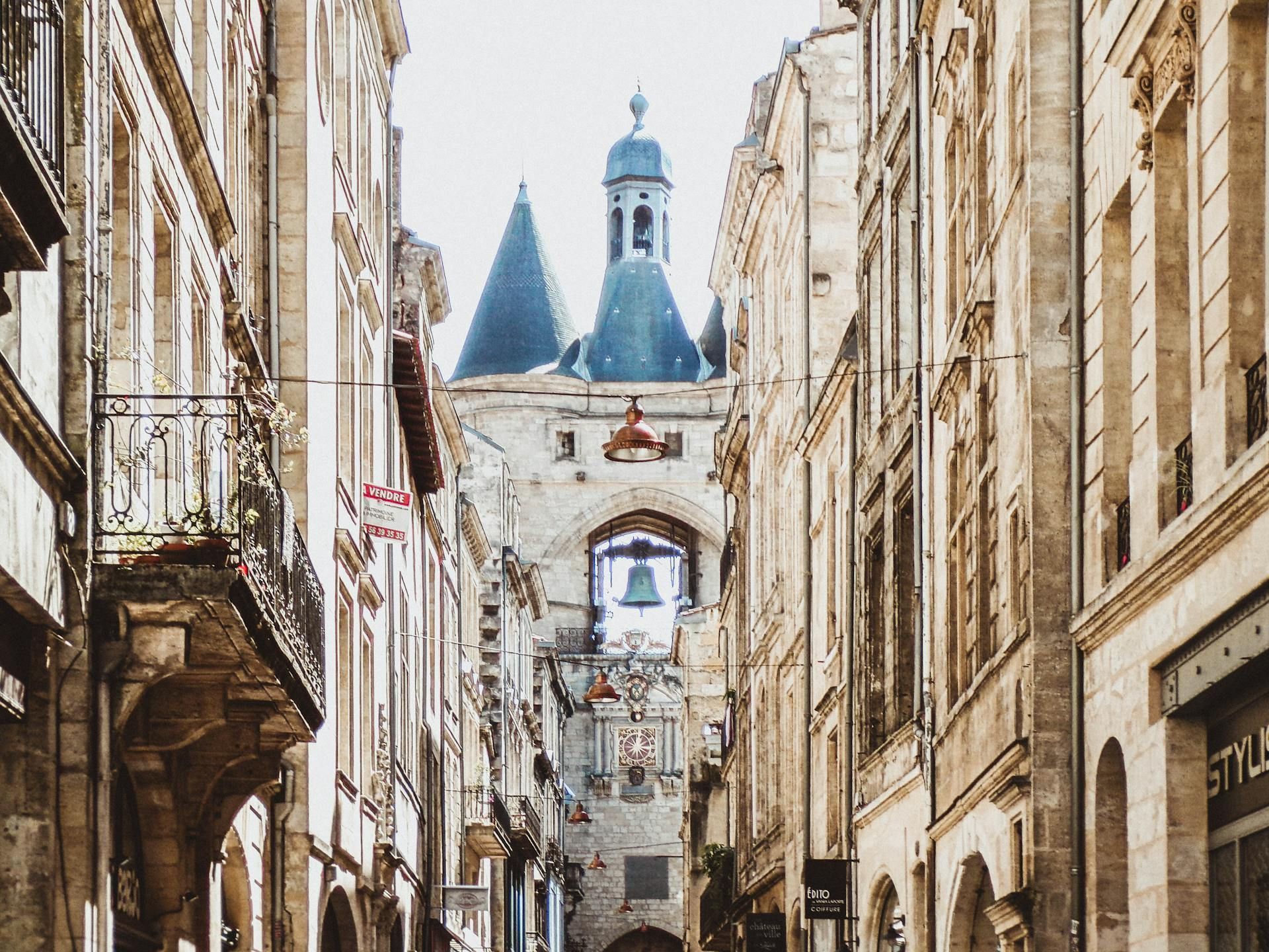 Una estrecha calle europea con edificios de piedra conduce a una caseta de vigilancia histórica con una gran campana visible bajo su arco.