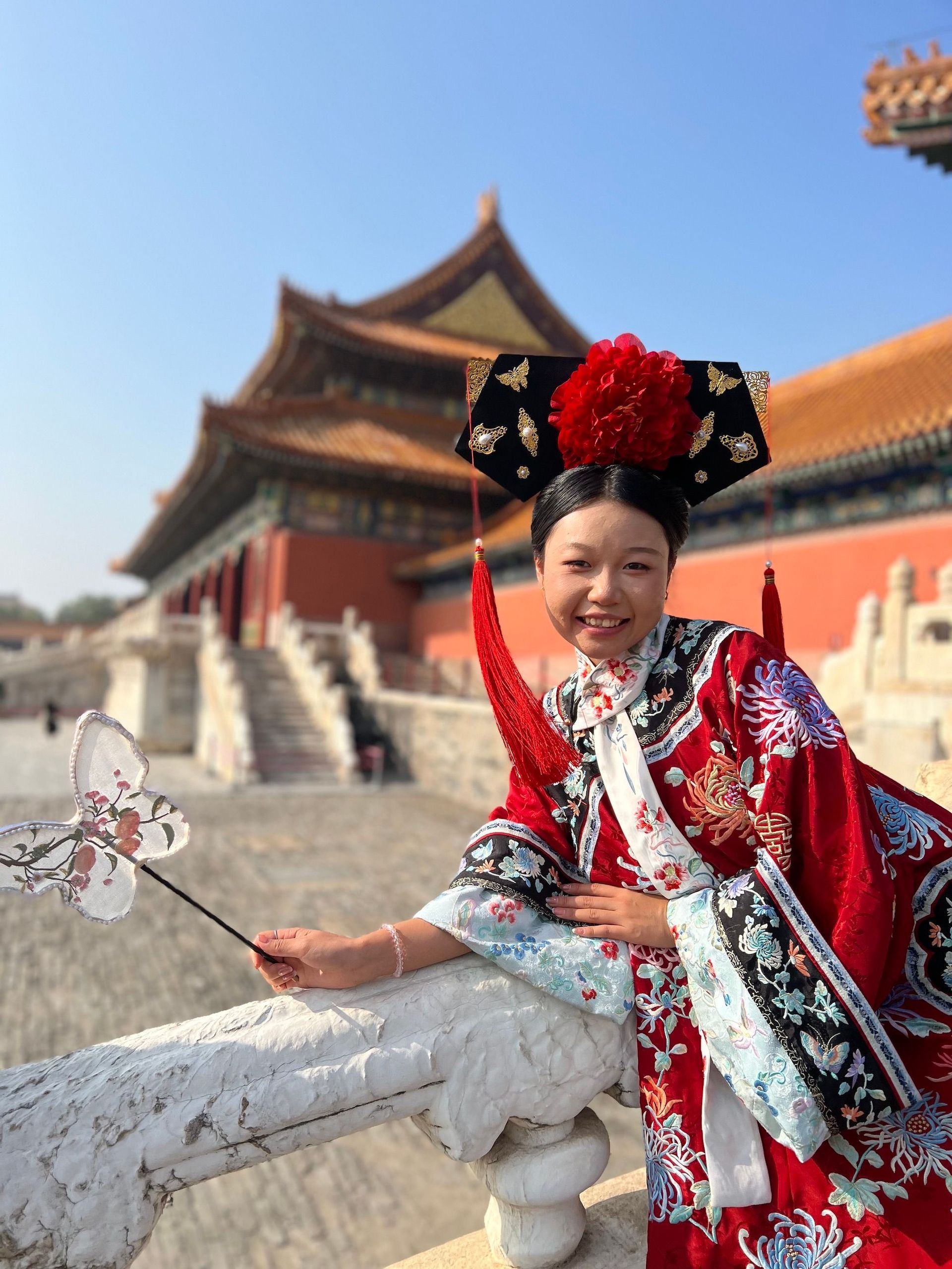 Una donna sorridente, in abito tradizionale rosso ricamato e copricapo, tiene in mano un ventaglio a forma di farfalla, con un palazzo cinese sullo sfondo.