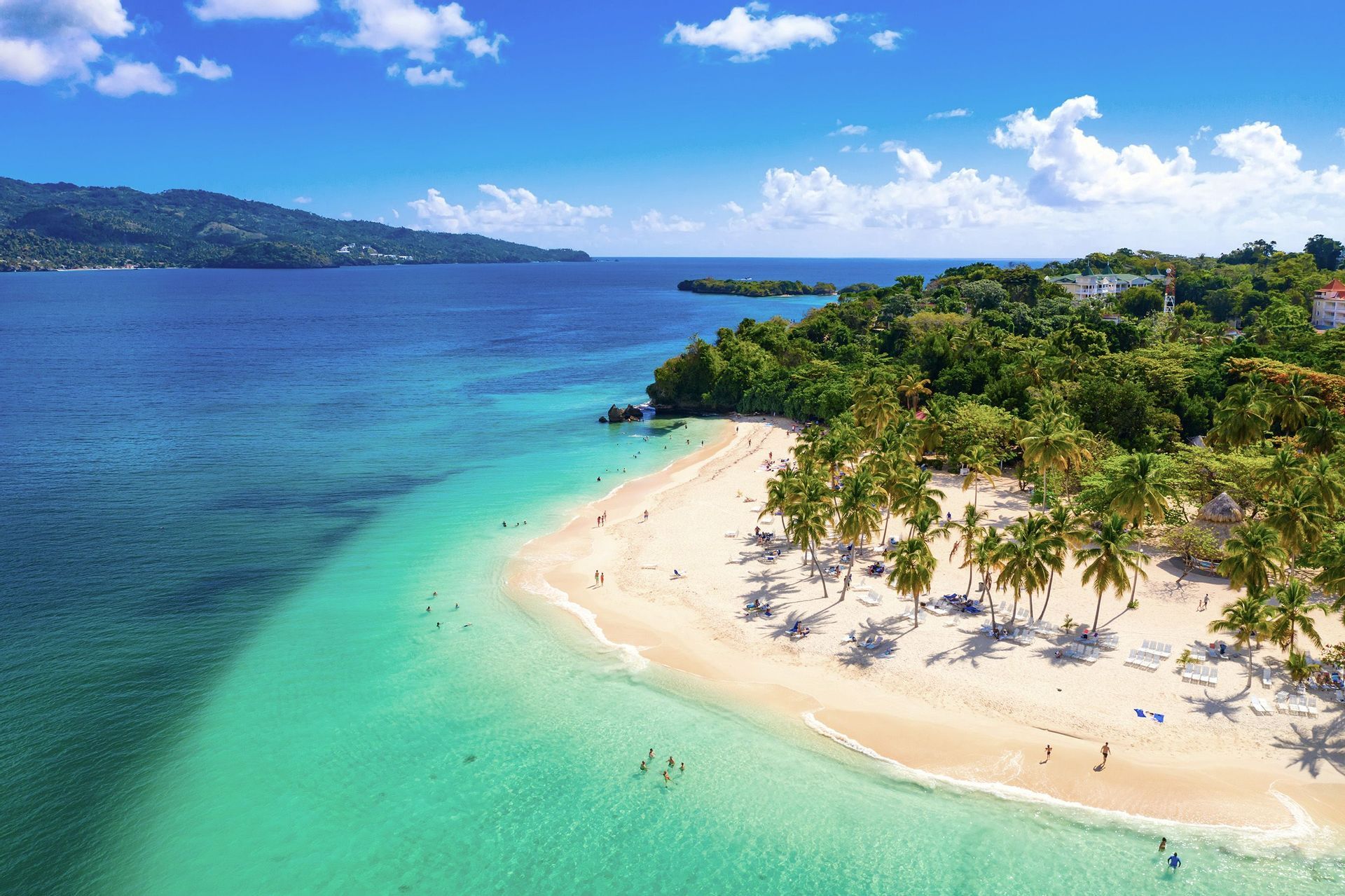 Veduta aerea di una spiaggia tropicale con persone che nuotano in acque turchesi e si rilassano su sabbia bianca tra palme lussureggianti.