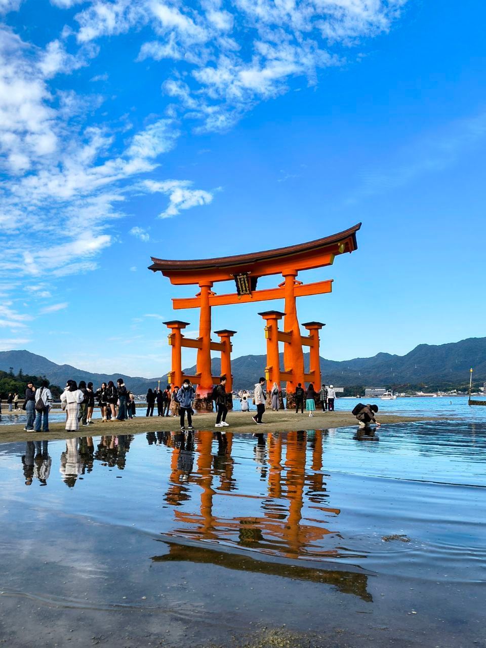 Un grande portale torii arancione si erge nell'acqua con la bassa marea, riflettendosi sulla superficie, mentre le persone si radunano sulla riva sotto un cielo azzurro.