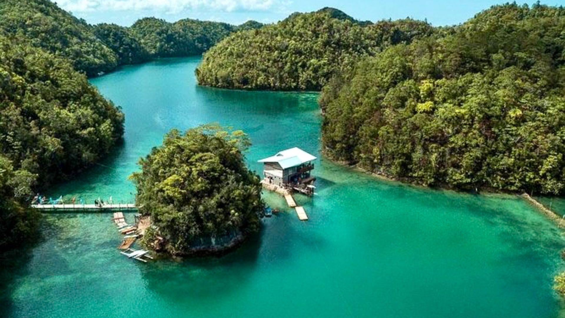 Una casa su palafitte su una piccola isola alberata in un fiume turchese, circondata da fitte colline verdi.