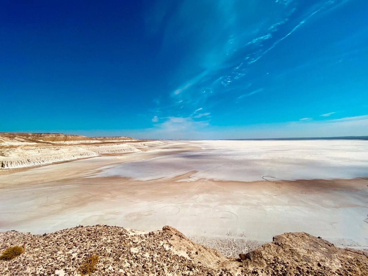 A panoramic view from a rocky outcrop over a vast, arid salt flat stretching to the horizon under a deep blue sky.