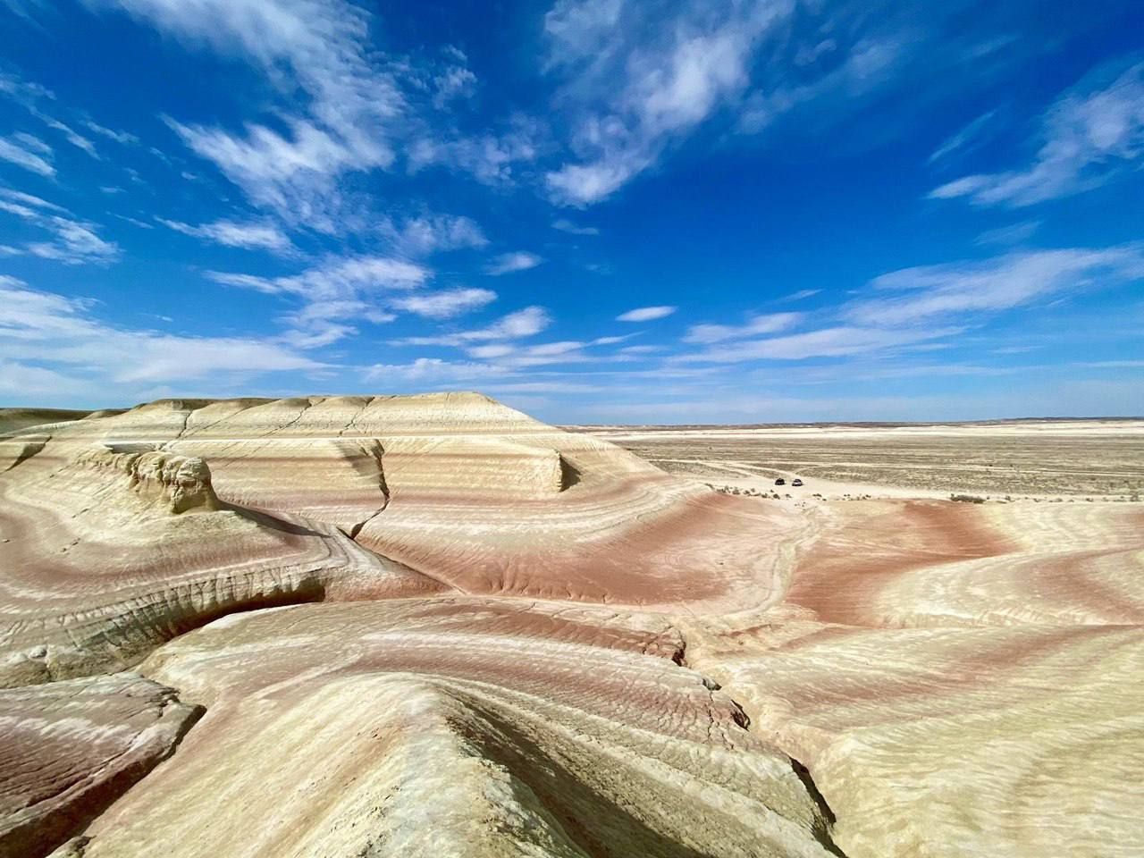 Formazioni rocciose multicolori e stratificate in un vasto paesaggio desertico sotto un cielo azzurro brillante con nuvole, con due veicoli in lontananza.