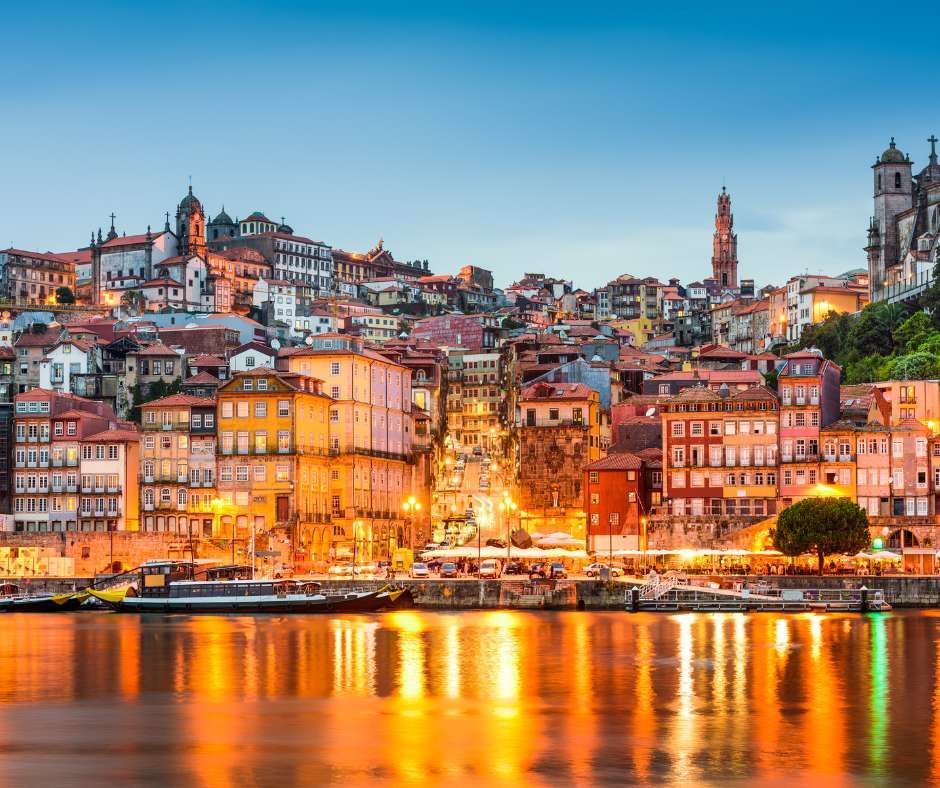 Eine farbenprächtige Stadtlandschaft am Hang, in der Abenddämmerung beleuchtet, mit Gebäudelichtern, die sich auf dem ruhigen Fluss spiegeln.