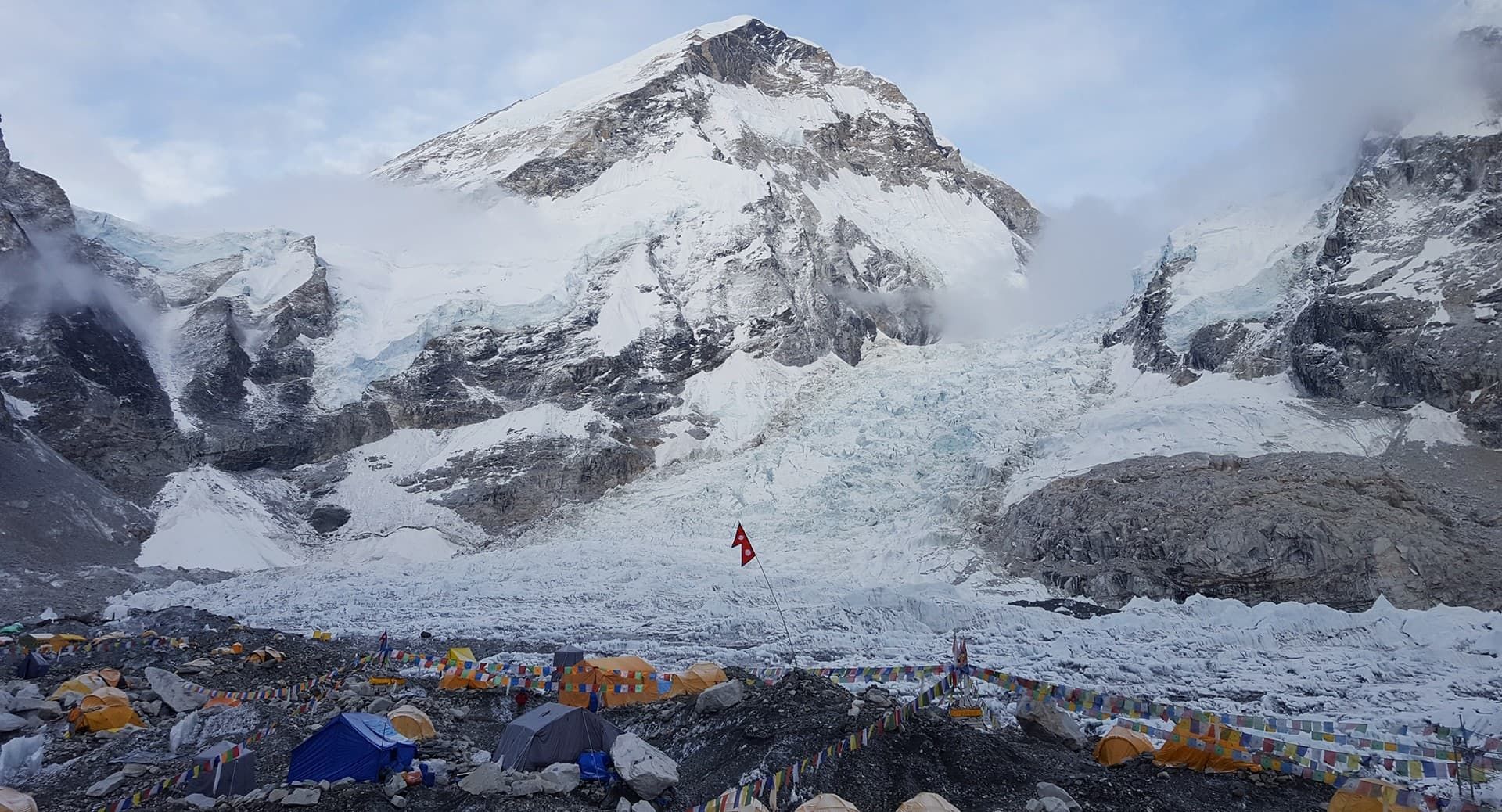 Un camp de base, avec ses tentes colorées et ses drapeaux de prière, est installé sur une moraine rocheuse, au pied d'une immense montagne enneigée et de son glacier.