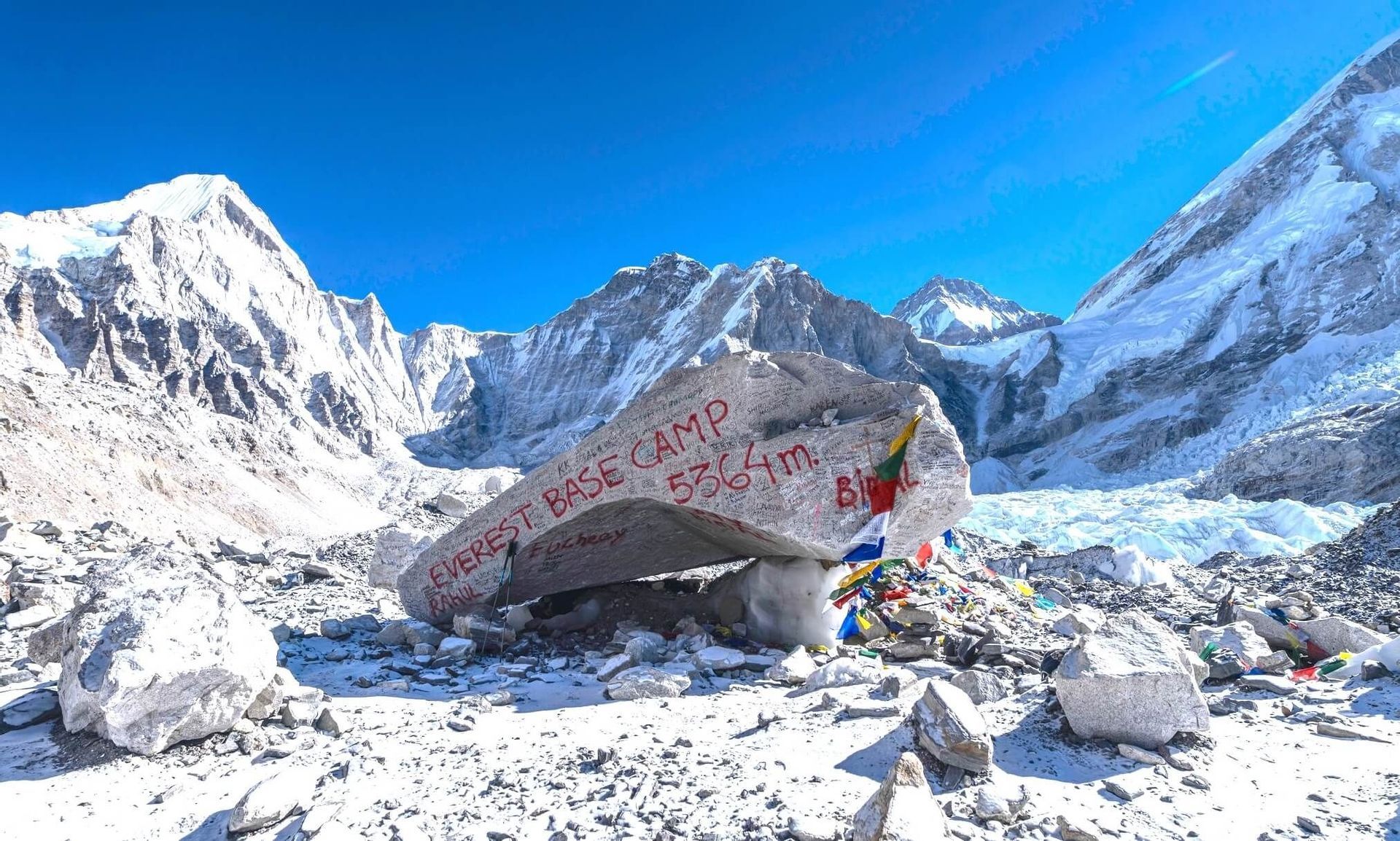 Der Markierungsfelsen des Everest-Basislagers, beschriftet mit seiner Höhe von 5364 m, befindet sich in einer verschneiten Landschaft, umgeben von Himalaya-Gipfeln unter klarem Himmel.