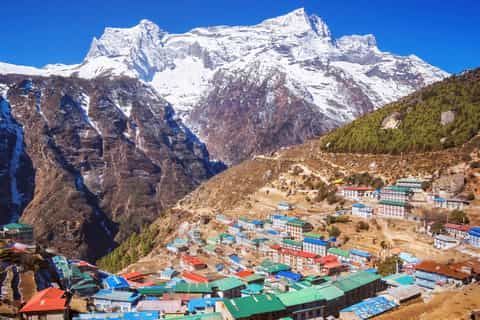 Un village aux toits colorés est niché à flanc de montagne escarpée, sous une grande montagne enneigée, sur fond de ciel bleu clair.