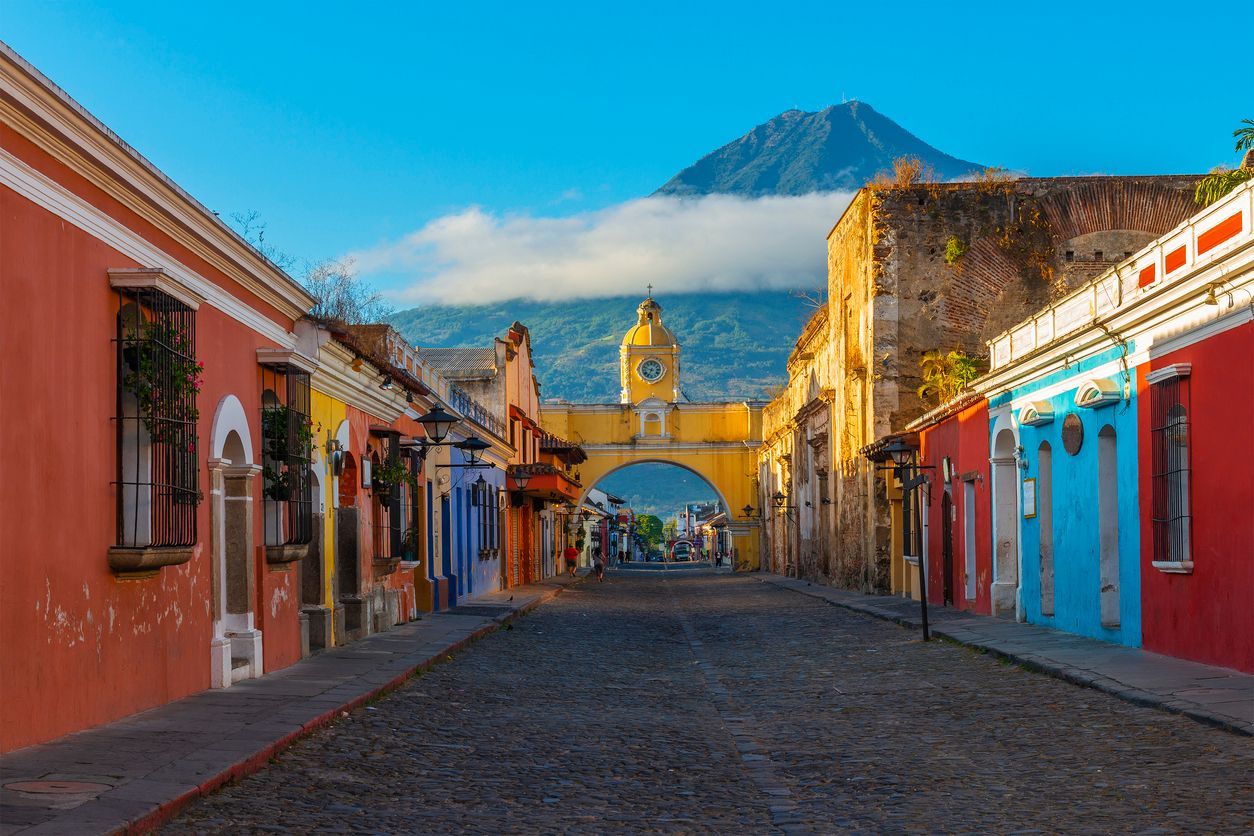 A cobblestone street lined with colorful colonial buildings leads to a yellow archway with a clock, with a volcano in the background under a blue sky.