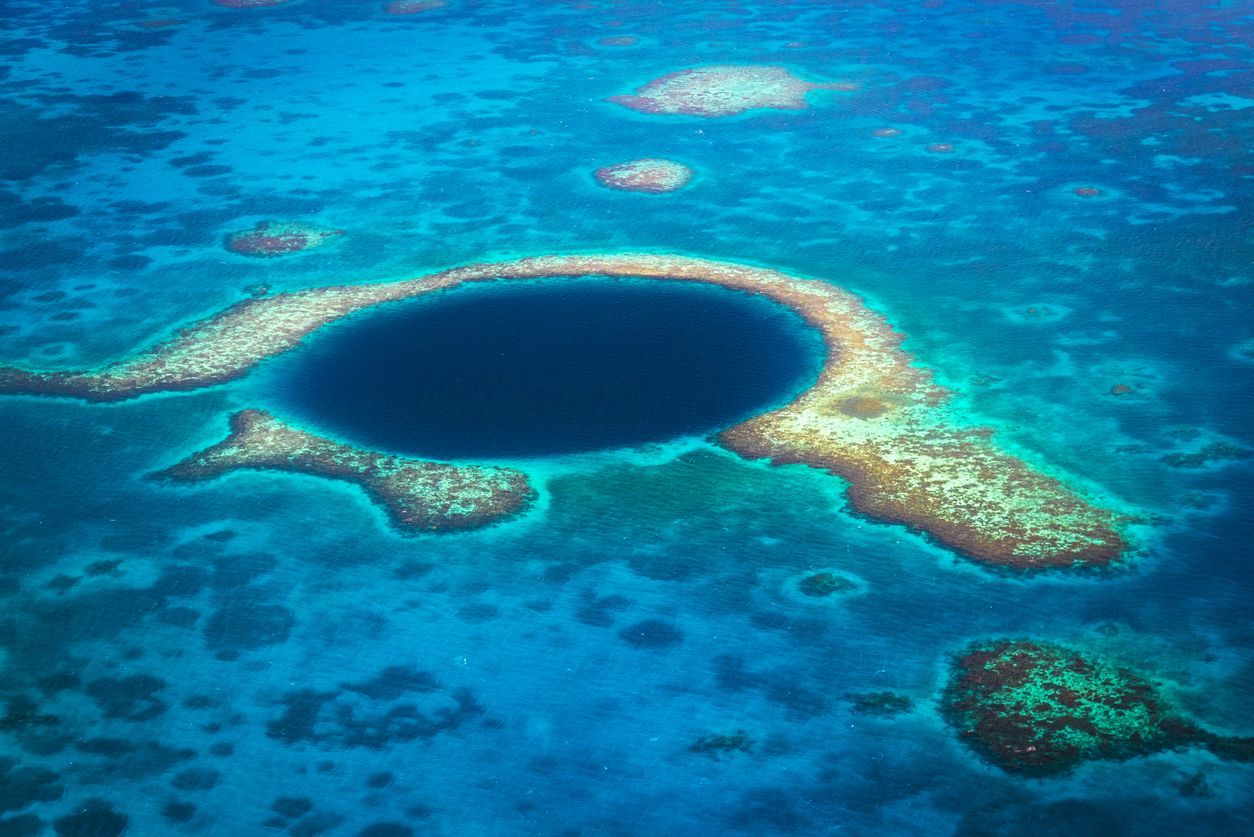 Une vue aérienne d'un grand gouffre marin bleu profond, entouré par un atoll de corail dans des eaux océaniques turquoises peu profondes.