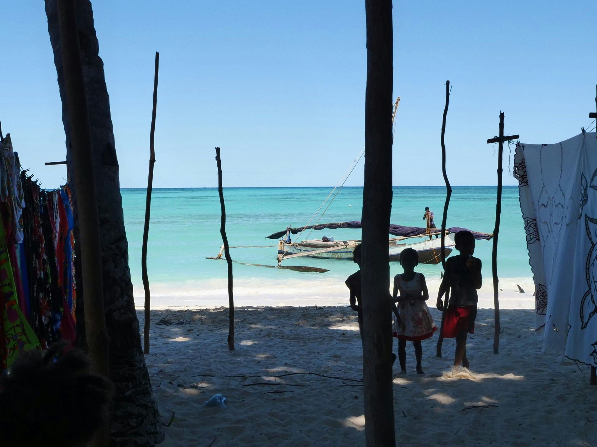 Trois enfants en silhouette se tiennent sur une plage de sable, regardant un bateau sur la mer turquoise sous un ciel bleu clair.