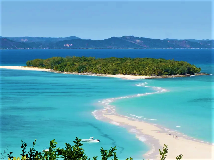 Un banc de sable blanc et sinueux s'étire d'une petite île arborée vers des eaux turquoise éclatantes, avec un bateau ancré à proximité.