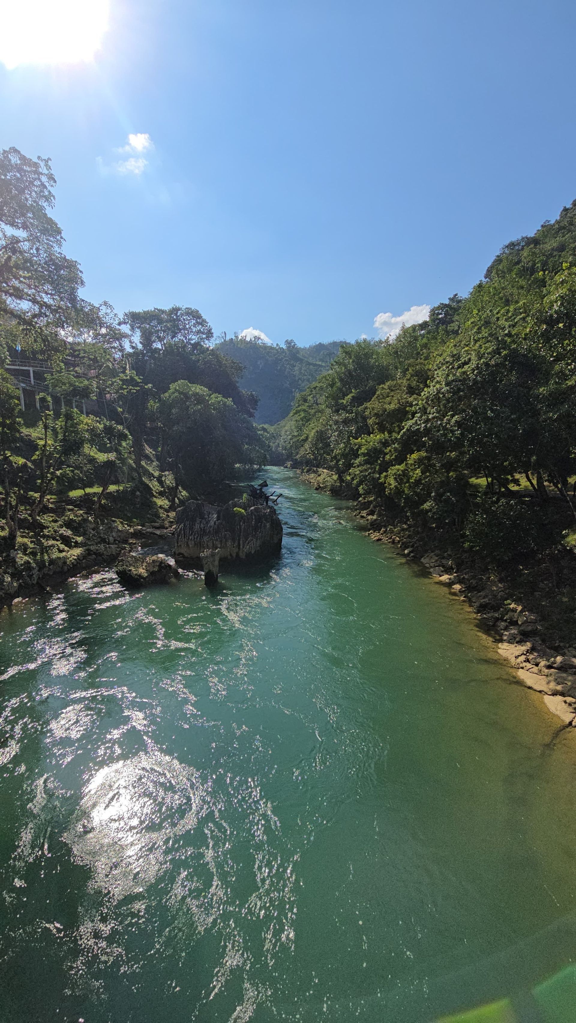 Un río turquesa fluye a través de un valle con exuberantes colinas verdes a ambos lados, bajo un cielo azul soleado.