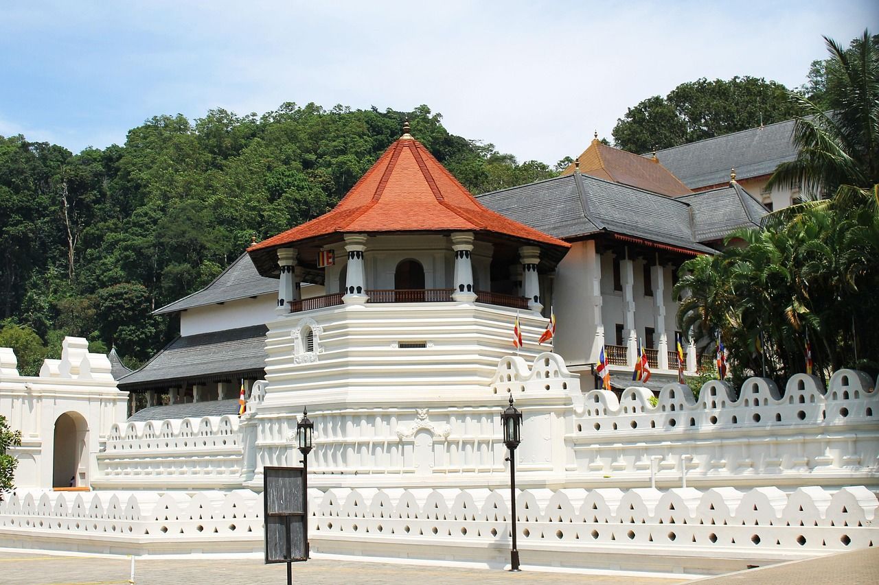 An ornate white temple complex with an octagonal, red-roofed tower set against a backdrop of a dense, green forest.