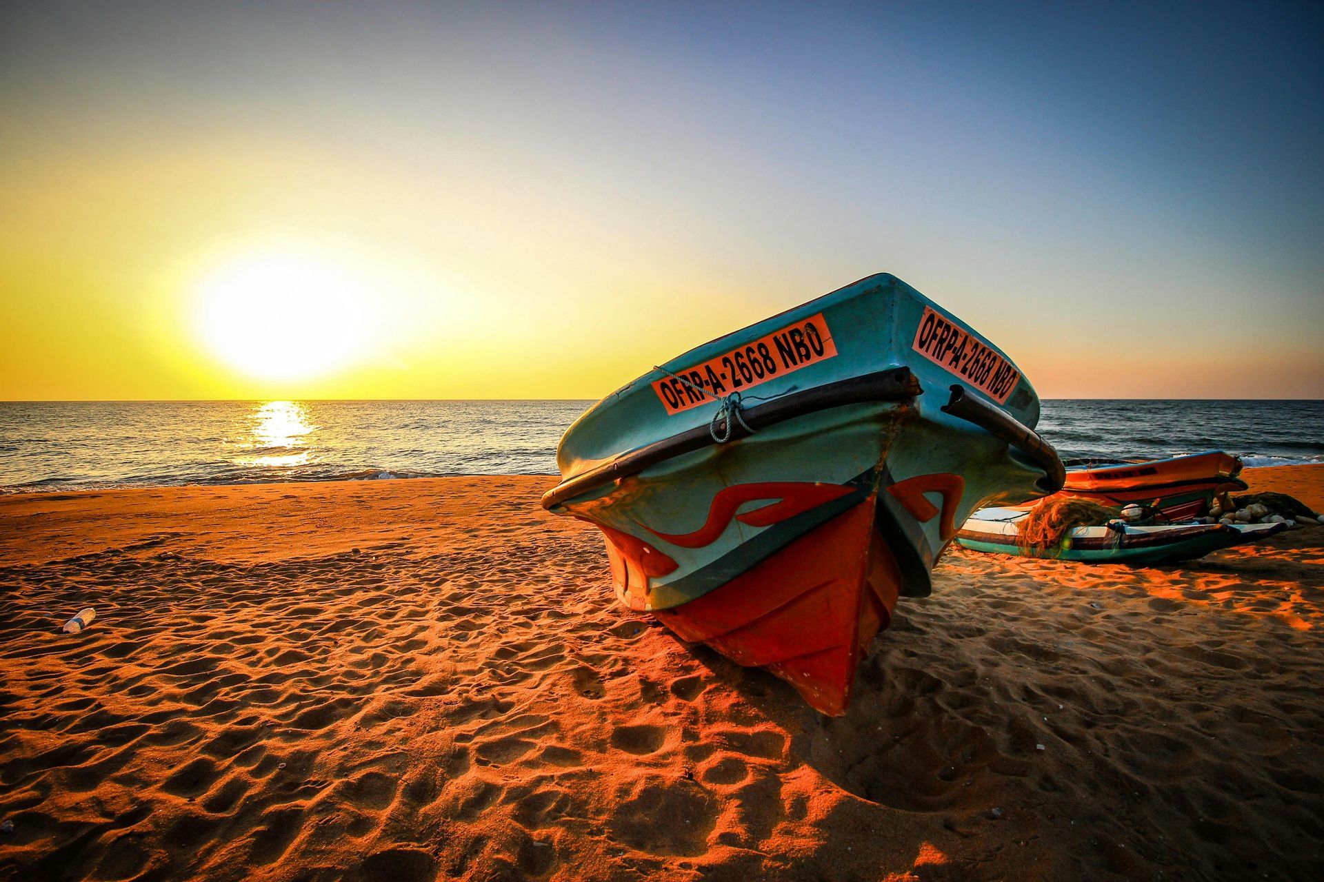 A colorful fishing boat rests on a sandy beach with the sun setting over the ocean, casting a golden glow.