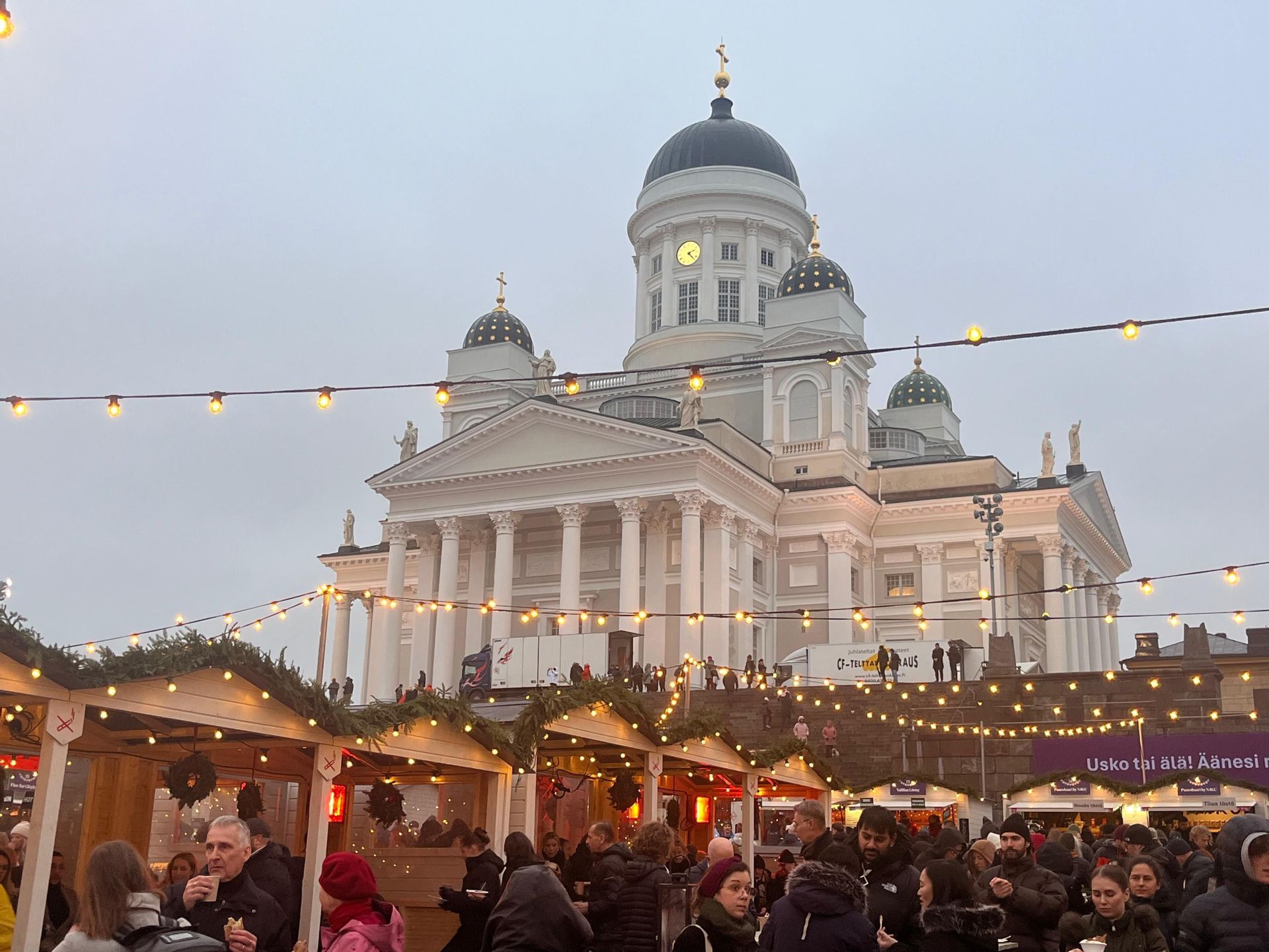 Un vivace mercatino di Natale con folla attorno a bancarelle in legno, di fronte a una grande cattedrale dalla cupola bianca sotto un cielo coperto.