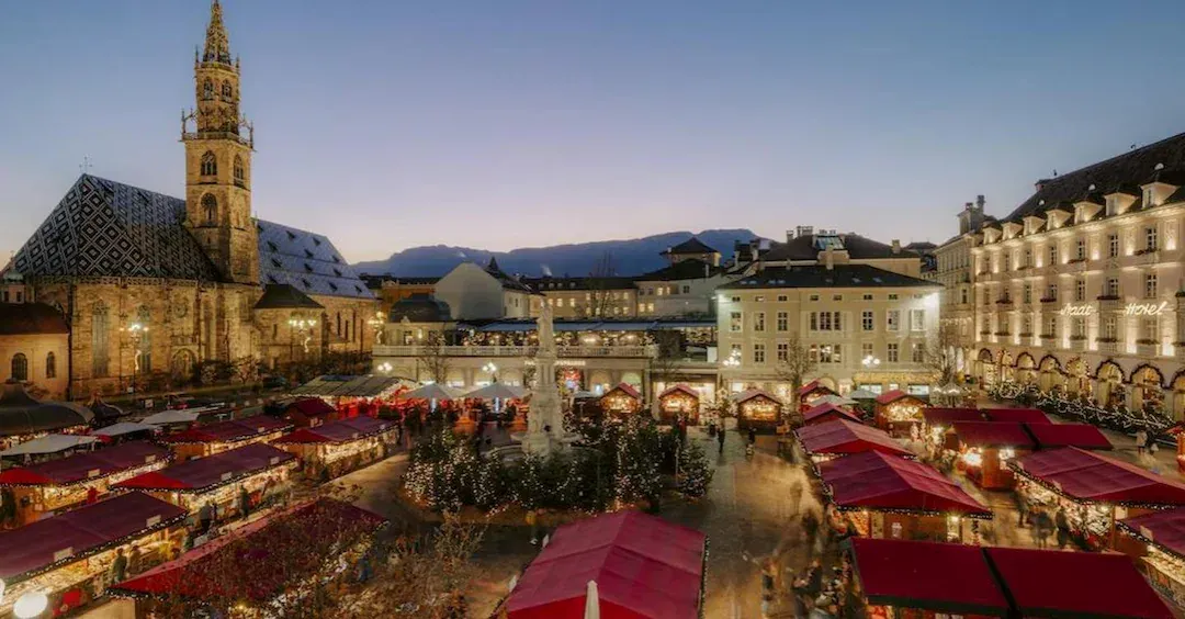 Una veduta aerea di un mercatino di Natale in una piazza del paese al crepuscolo, con bancarelle illuminate accanto a una grande chiesa gotica.