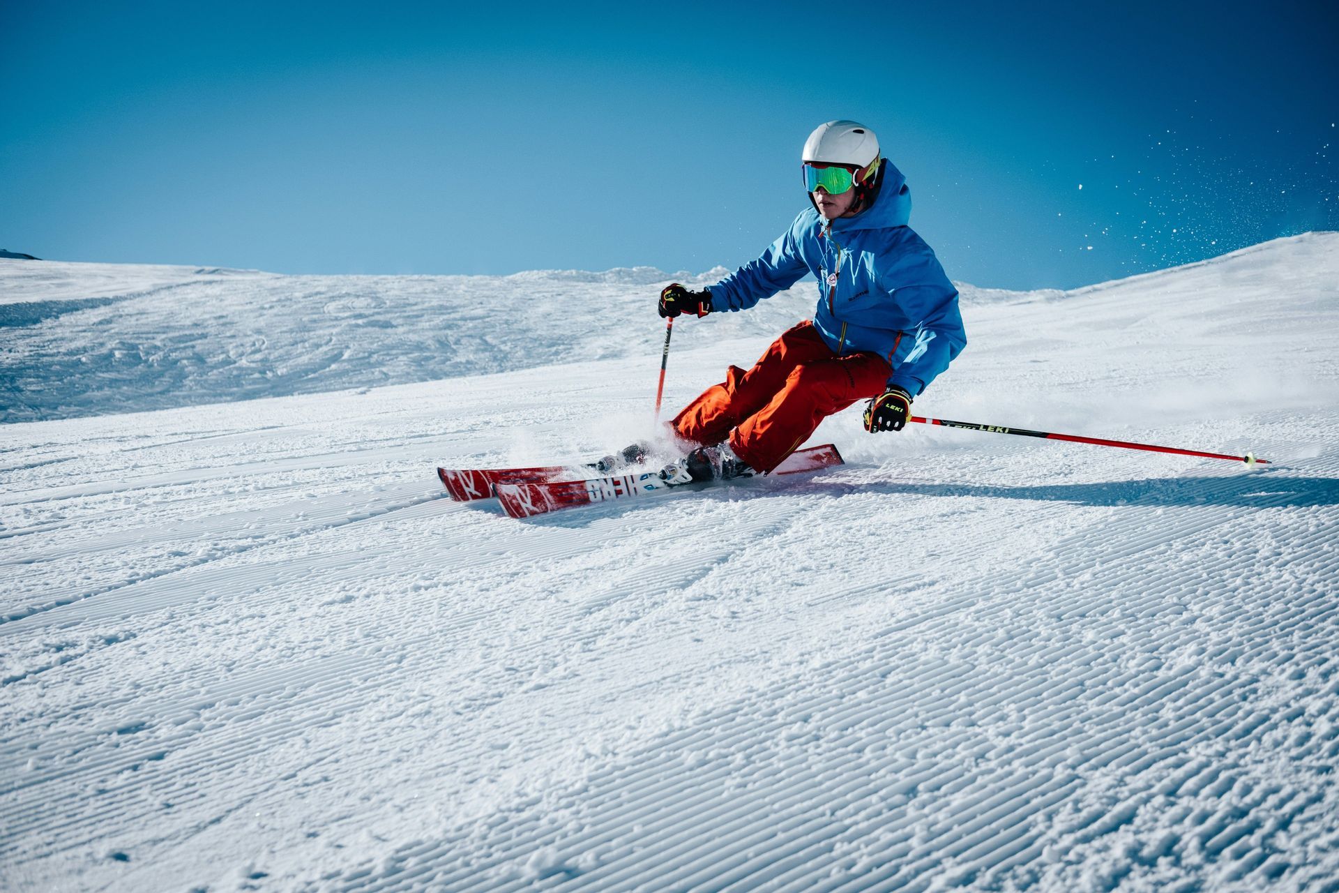 Una persona con giacca blu e pantaloni arancioni scia su una pista innevata battuta sotto un cielo azzurro limpido.