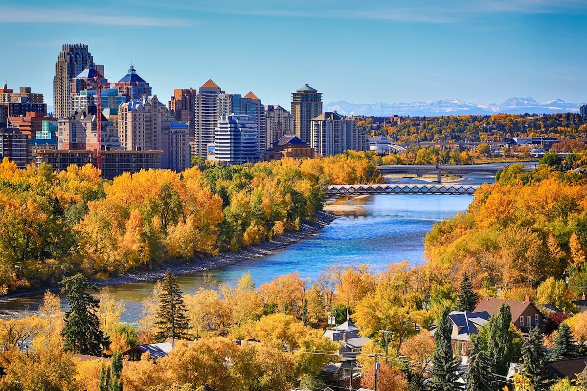 A city skyline overlooks a winding river, its banks lined with golden autumn trees, with distant mountains under a blue sky.