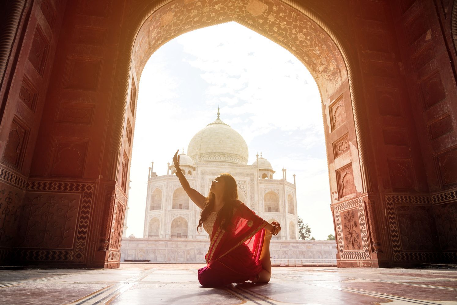 Una donna in un sari rosso pratica yoga sotto un arco di pietra, che incornicia la vista di un mausoleo a cupola di marmo bianco.