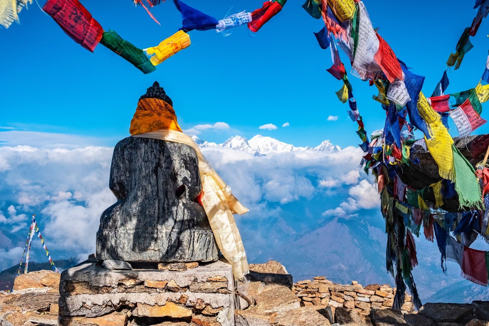 Una estatua de piedra vista desde atrás contempla una cordillera nevada y un mar de nubes, bajo coloridas banderas de oración tibetanas.