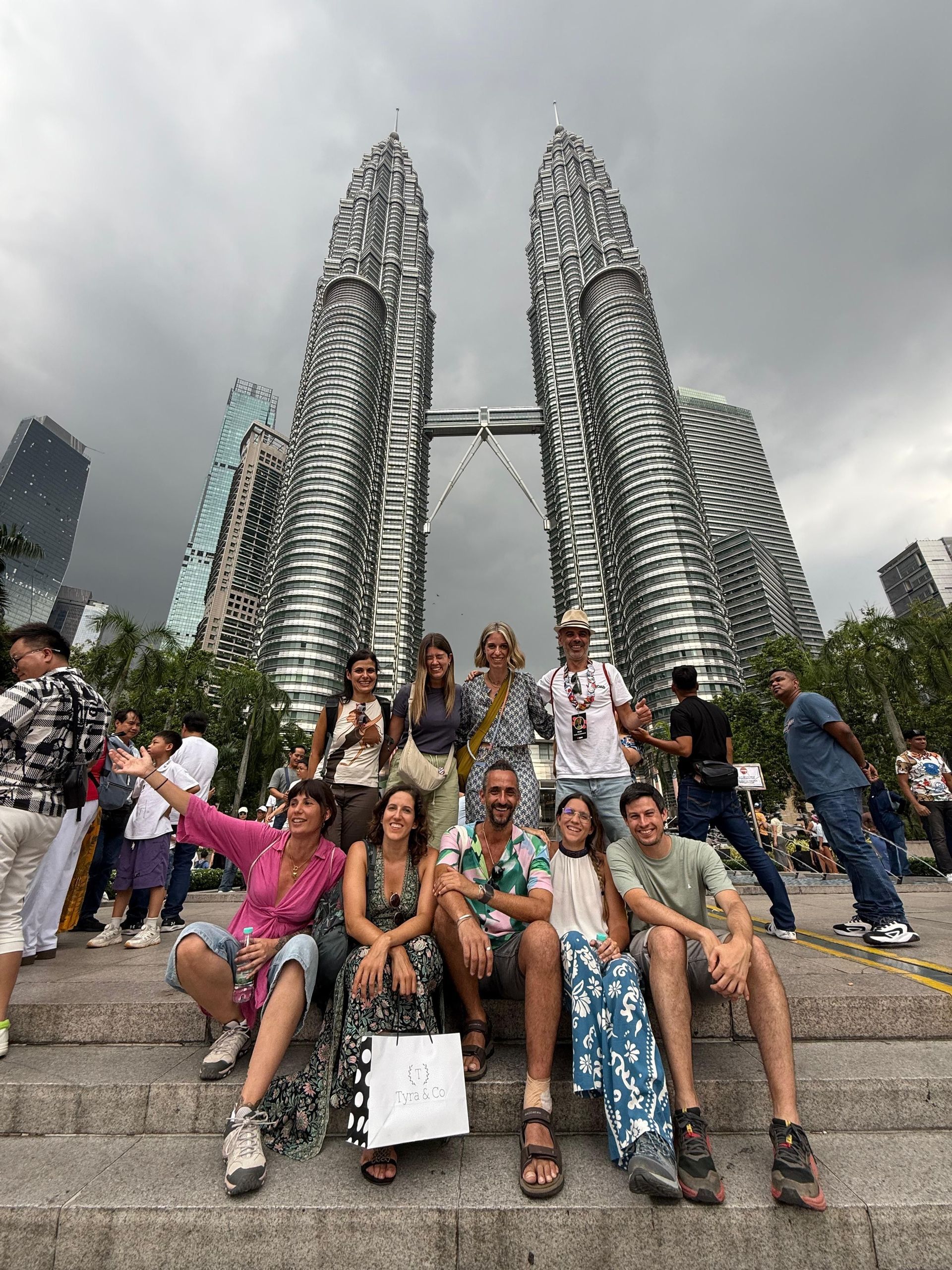 Eine WeRoad Gruppenreise posiert für ein Foto auf Stufen vor hohen Zwillingswolkenkratzern unter einem bewölkten Himmel.