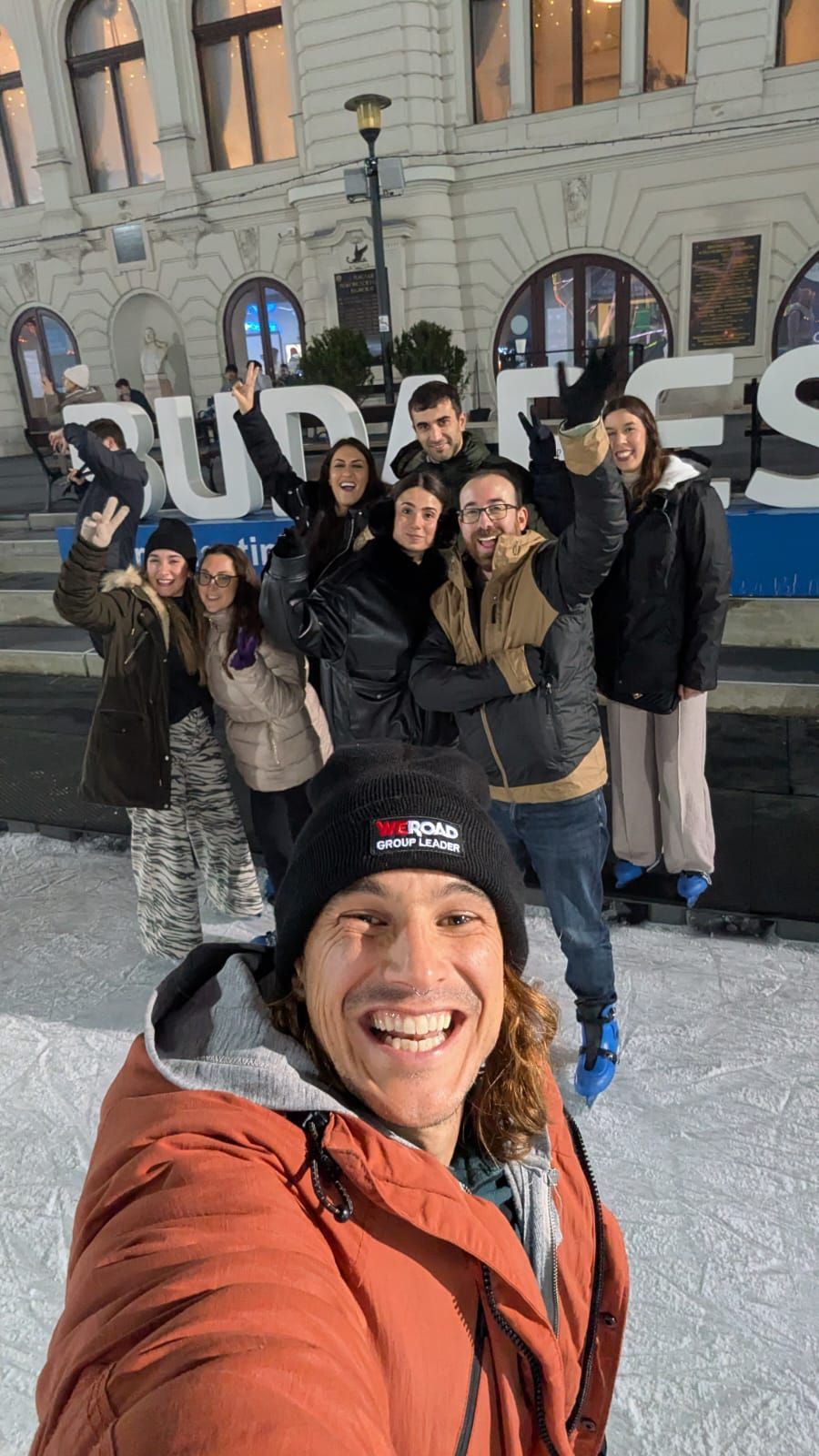 Un capogruppo WeRoad si scatta un selfie sorridente con il suo gruppo mentre pattinano sul ghiaccio all'aperto di fronte a un grande edificio.