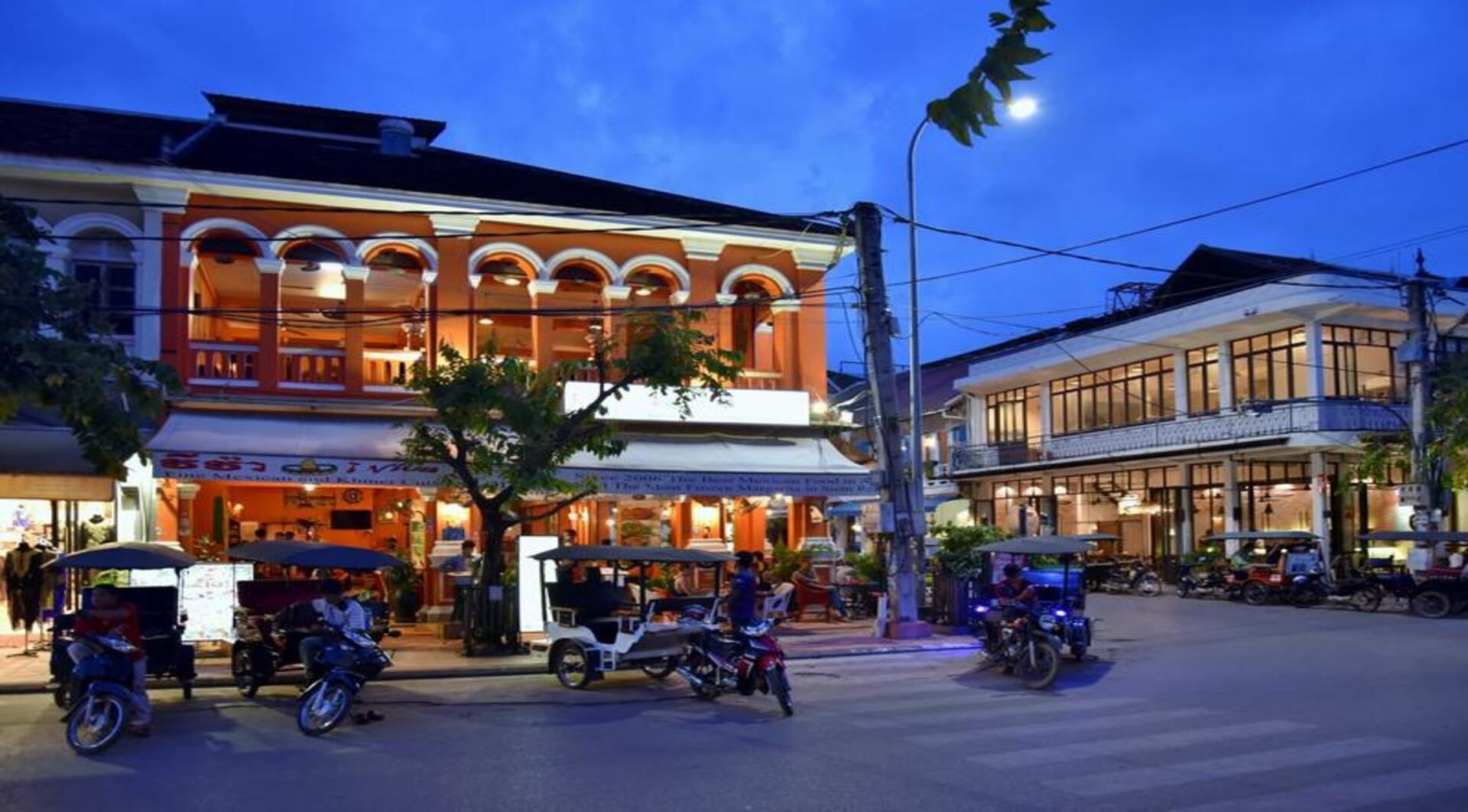 Tuk-tuk e scooter sono parcheggiati all'angolo di una strada cittadina davanti a edifici in stile coloniale illuminati al tramonto.