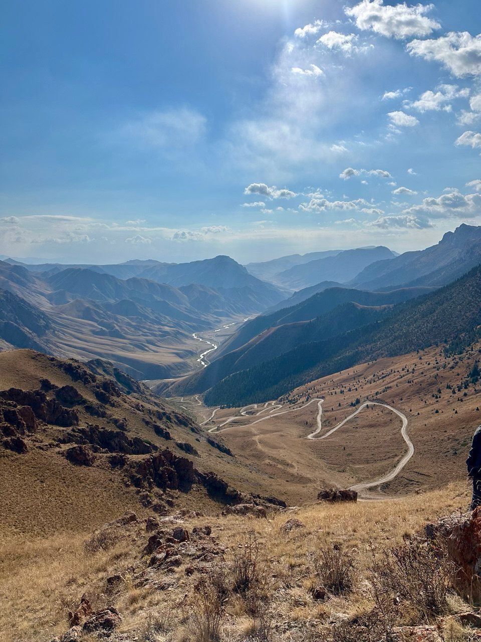 Une vue plongeante d'une vaste vallée montagneuse, avec une route sinueuse et une rivière qui serpente à travers le paysage, sous un ciel bleu et ensoleillé parsemé de légers nuages.