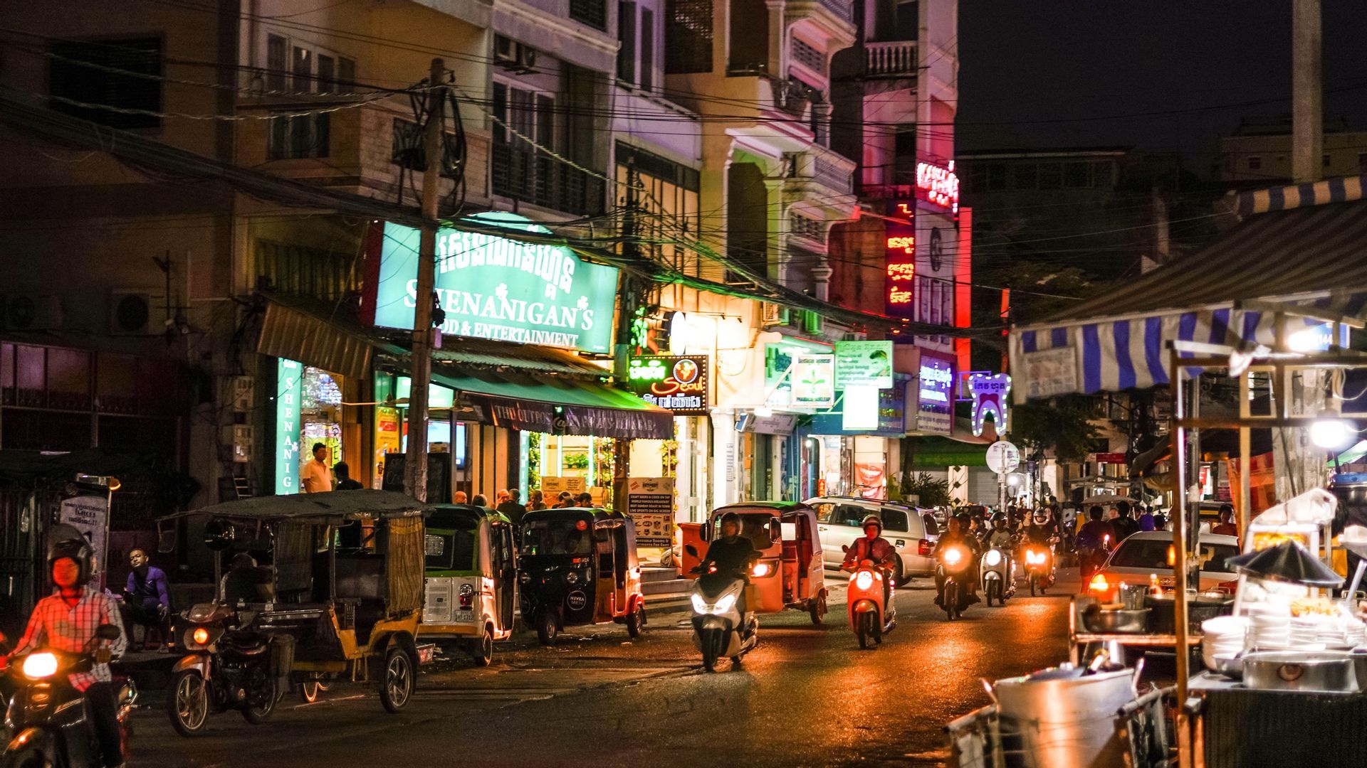 A busy city street at night with traffic from motorcycles and tuk-tuks under colorful neon signs.