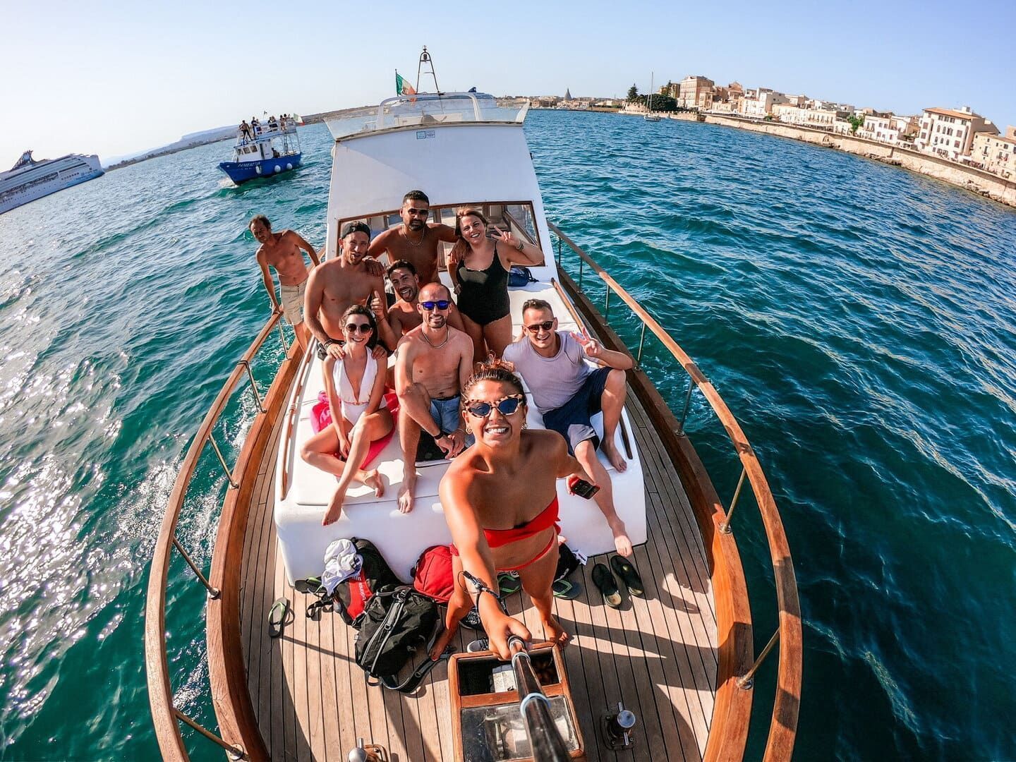 Un gruppo WeRoad si scatta un selfie sul ponte di legno di una barca che naviga sul mare, con una città costiera sullo sfondo.