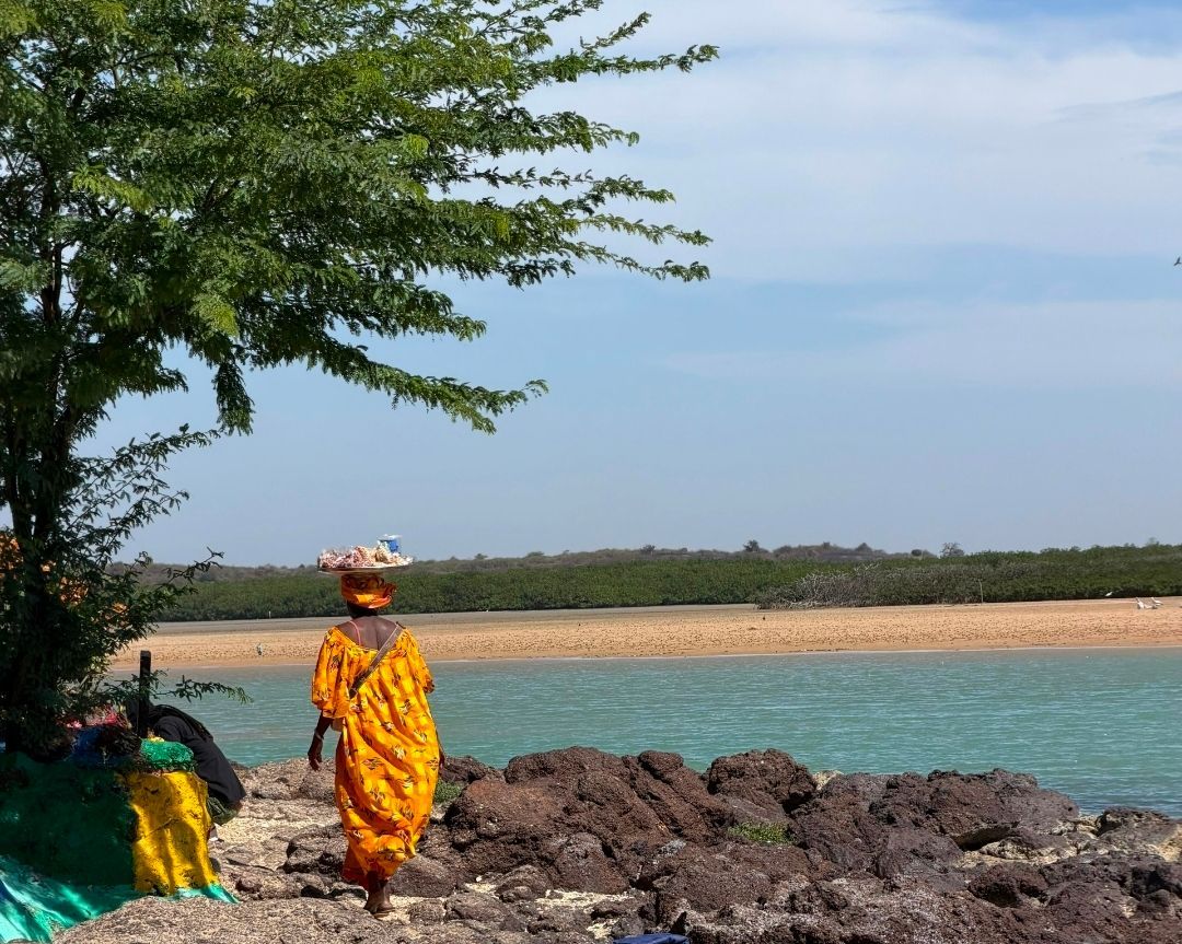 Una mujer con un vibrante vestido naranja camina por una costa rocosa, equilibrando una bandeja de productos en su cabeza junto a aguas turquesas.