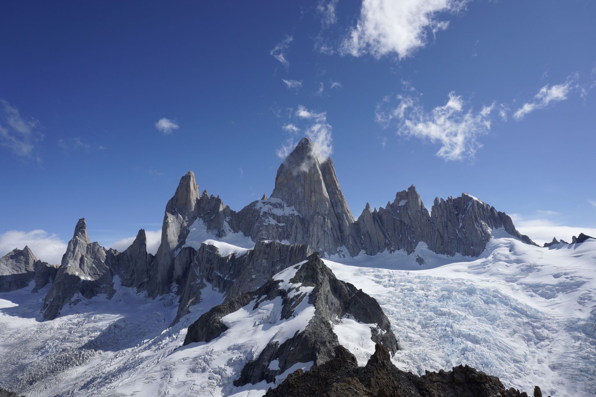 A range of jagged, rocky mountains covered in snow and glaciers under a bright blue sky with sparse clouds.
