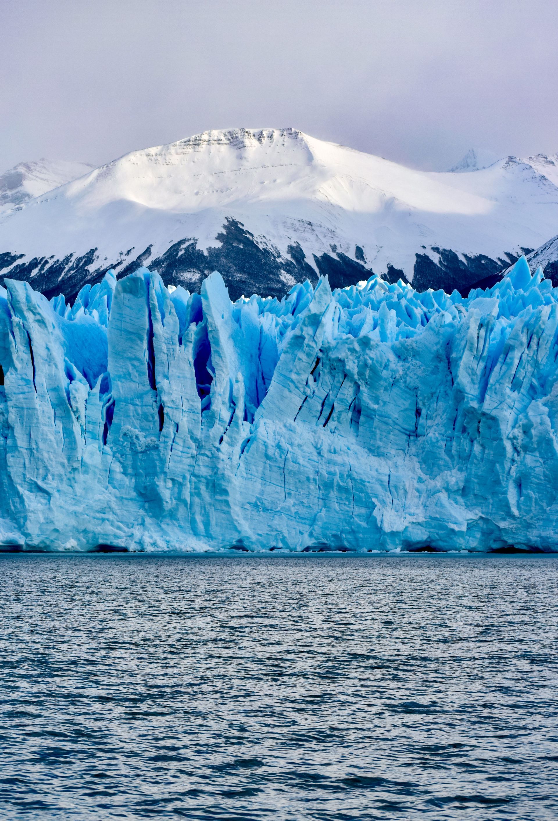 Un immense glacier bleu s'élève de l'eau sombre, avec des montagnes enneigées en arrière-plan.