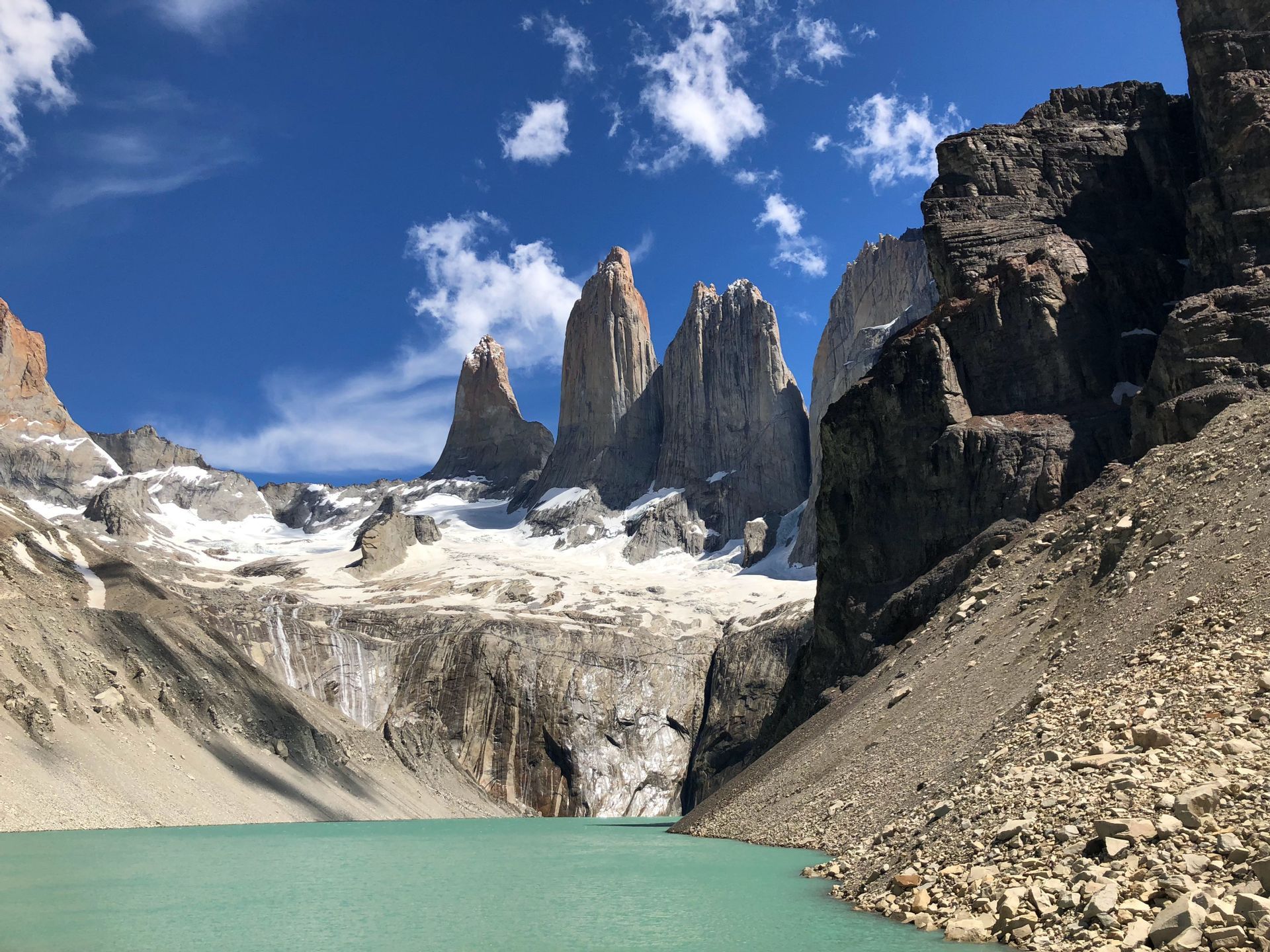 Tre cime granitiche frastagliate e un ghiacciaio che svetta su un lago glaciale turchese sotto un cielo azzurro brillante con nuvole sparse.