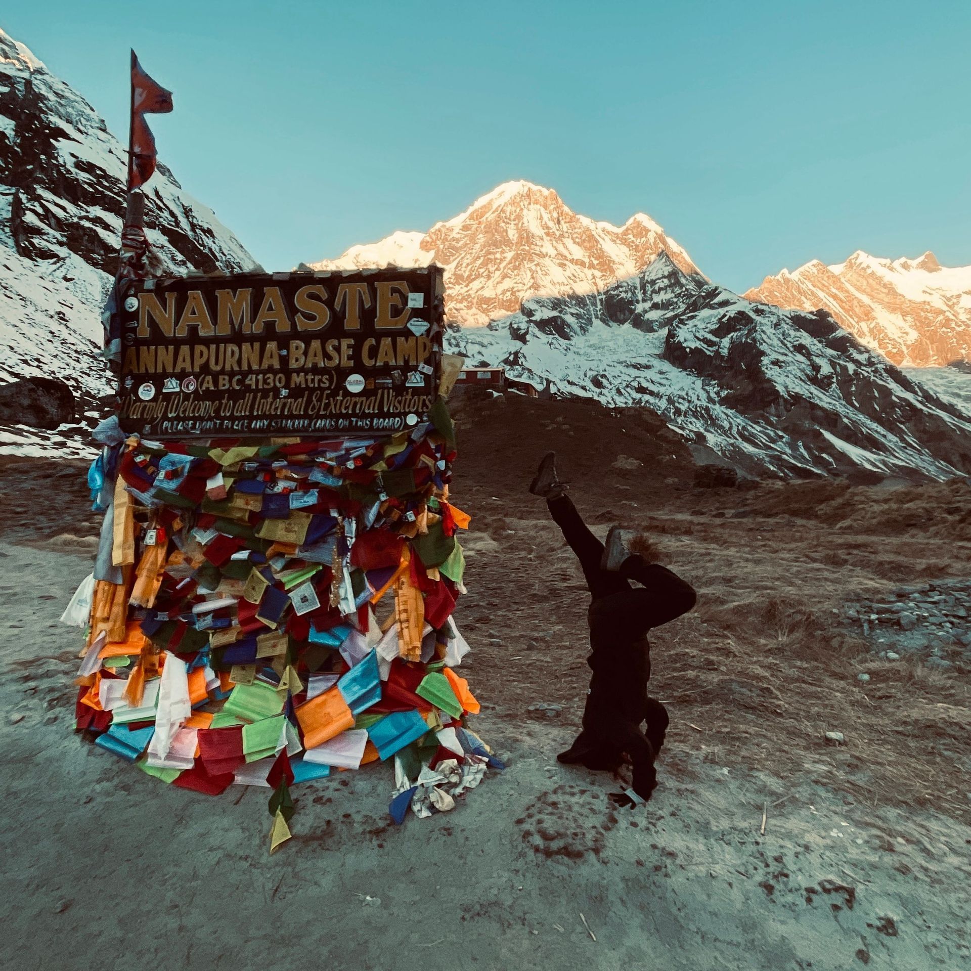 Una persona hace una parada de cabeza junto al letrero del Campamento Base Annapurna, con montañas nevadas e iluminadas por el sol de fondo.