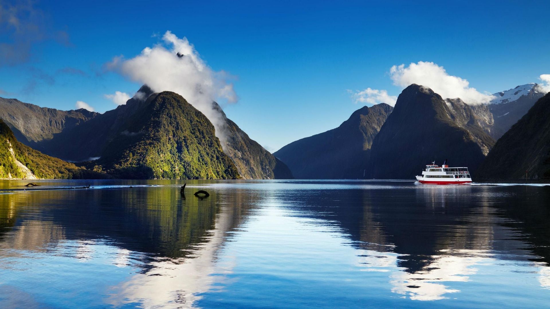A red and white tour boat sails on calm water through a fjord, with steep, green mountains reflected on the surface.