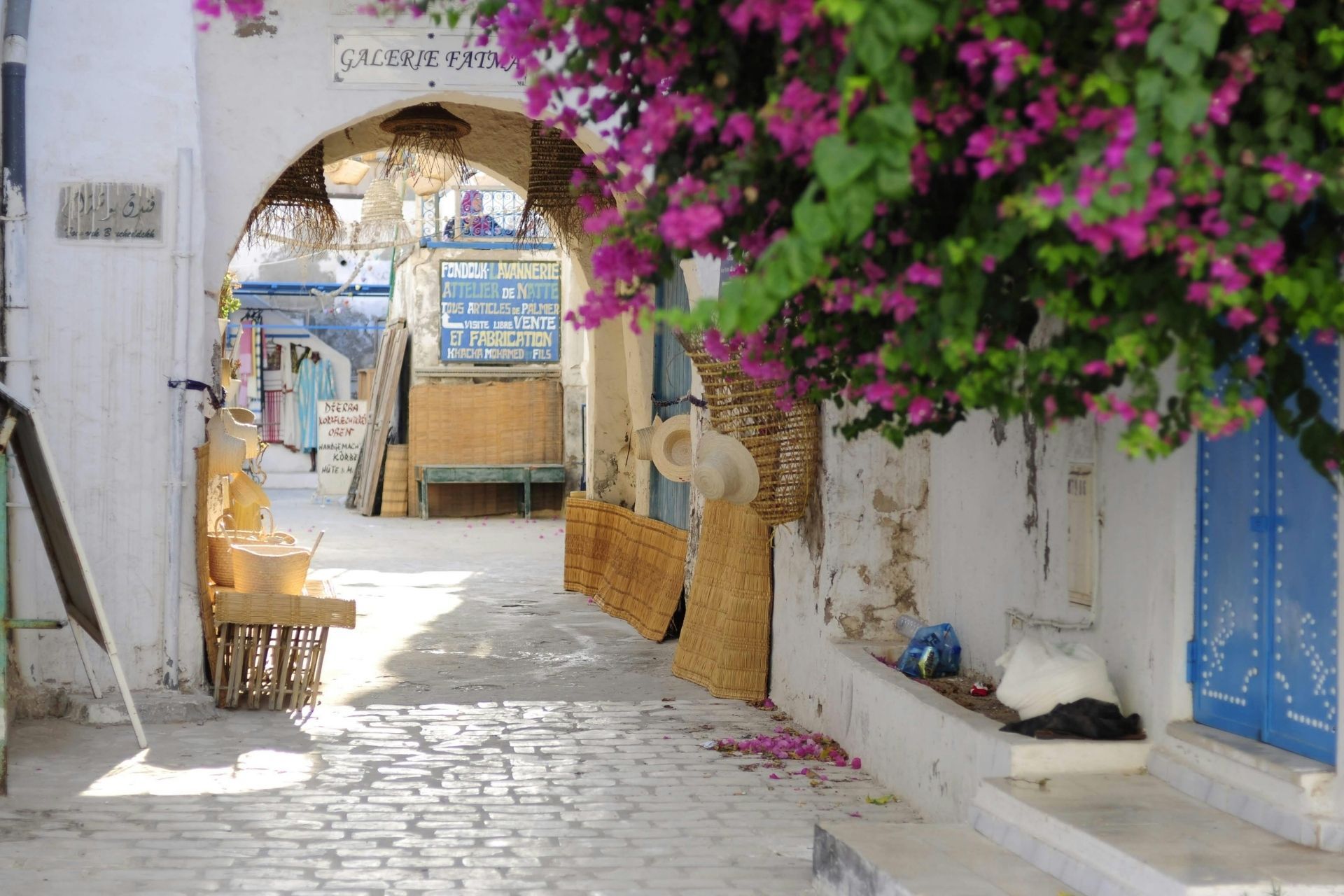 Eine sonnenbeschienene Kopfsteinpflastergasse auf einem Markt, mit Kunsthandwerk zum Verkauf und leuchtend rosa Bougainvillea-Blüten, die darüber hängen.