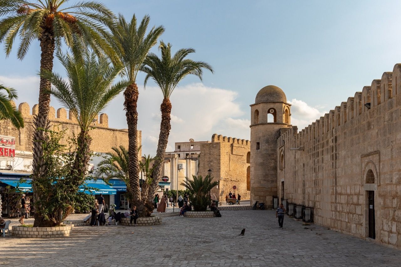 Ein sonniger Kopfsteinpflasterplatz mit Palmen, wo sich Menschen nahe alter Steinfestungen und eines Kuppelturms versammeln.