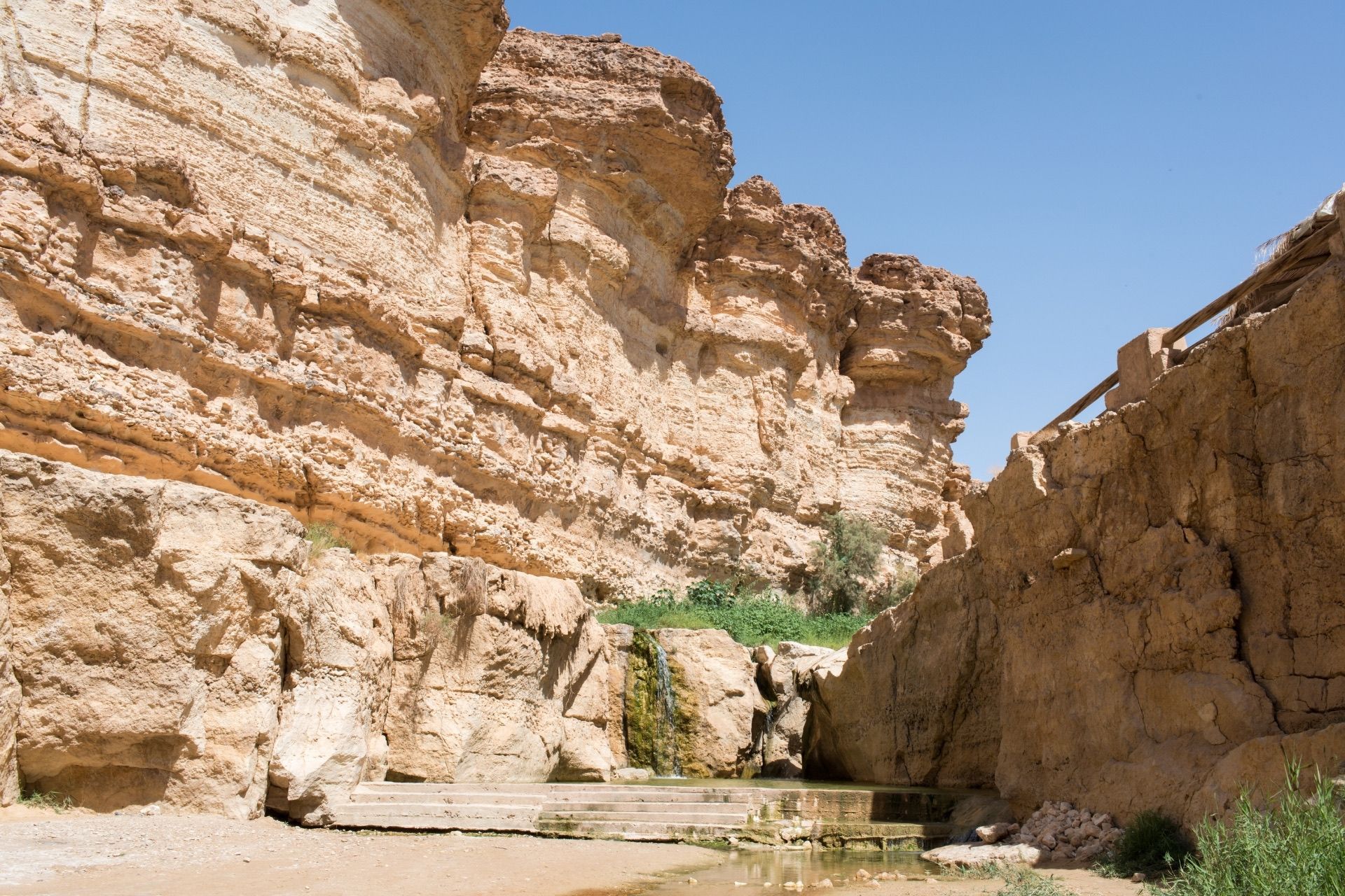 Am Fuße einer geschichteten Felsenschlucht stürzt ein kleiner Wasserfall in ein Wasserbecken, zu dem Steinstufen hinabführen.