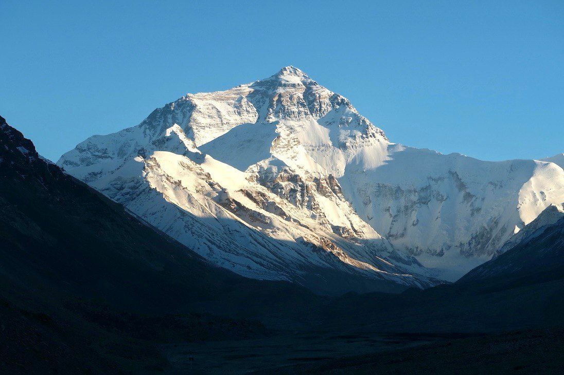 Una grande cima montuosa coperta di neve è illuminata dal sole contro un cielo azzurro limpido, con una valle scura e in ombra in primo piano.
