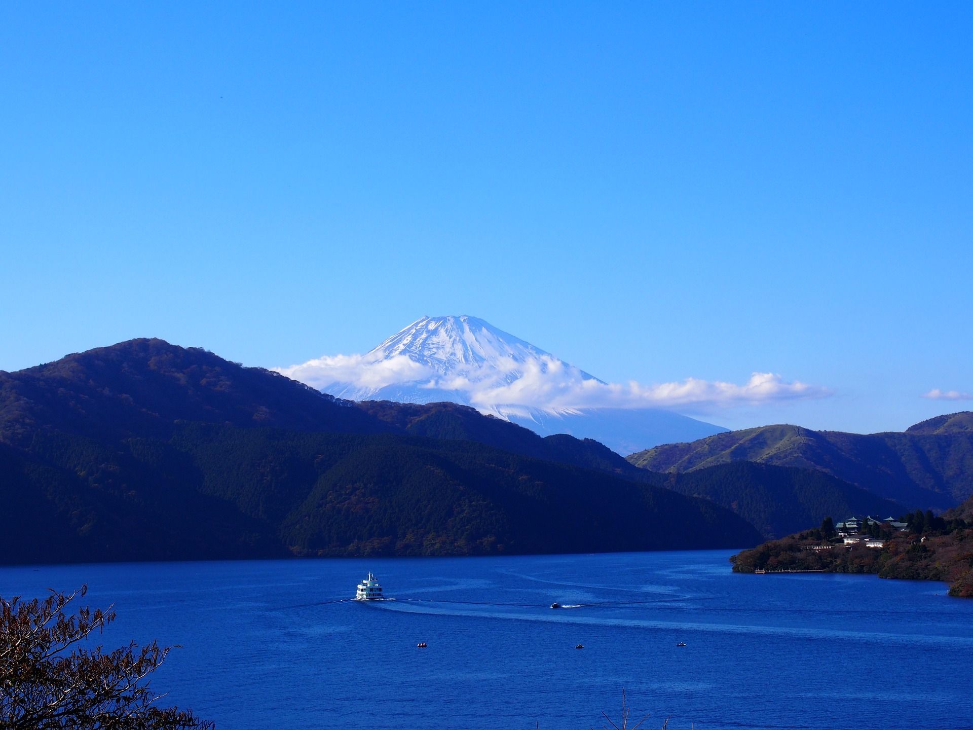 Un traghetto bianco naviga su un lago azzurro, con montagne boscose e il Monte Fuji innevato visibili sullo sfondo.